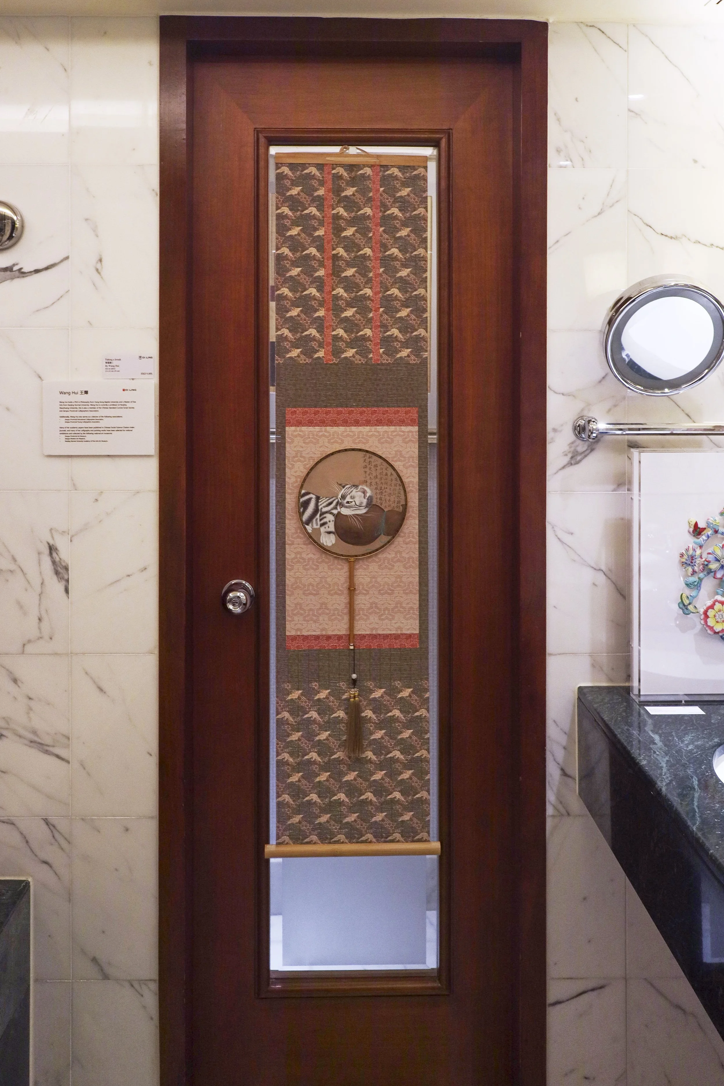 Whatz Art Fair 2025: Decorative hanging with a round frame showing a cat playing with a bowl, surrounded by intricate patterned fabric in shades of red, pink, and dark brown.