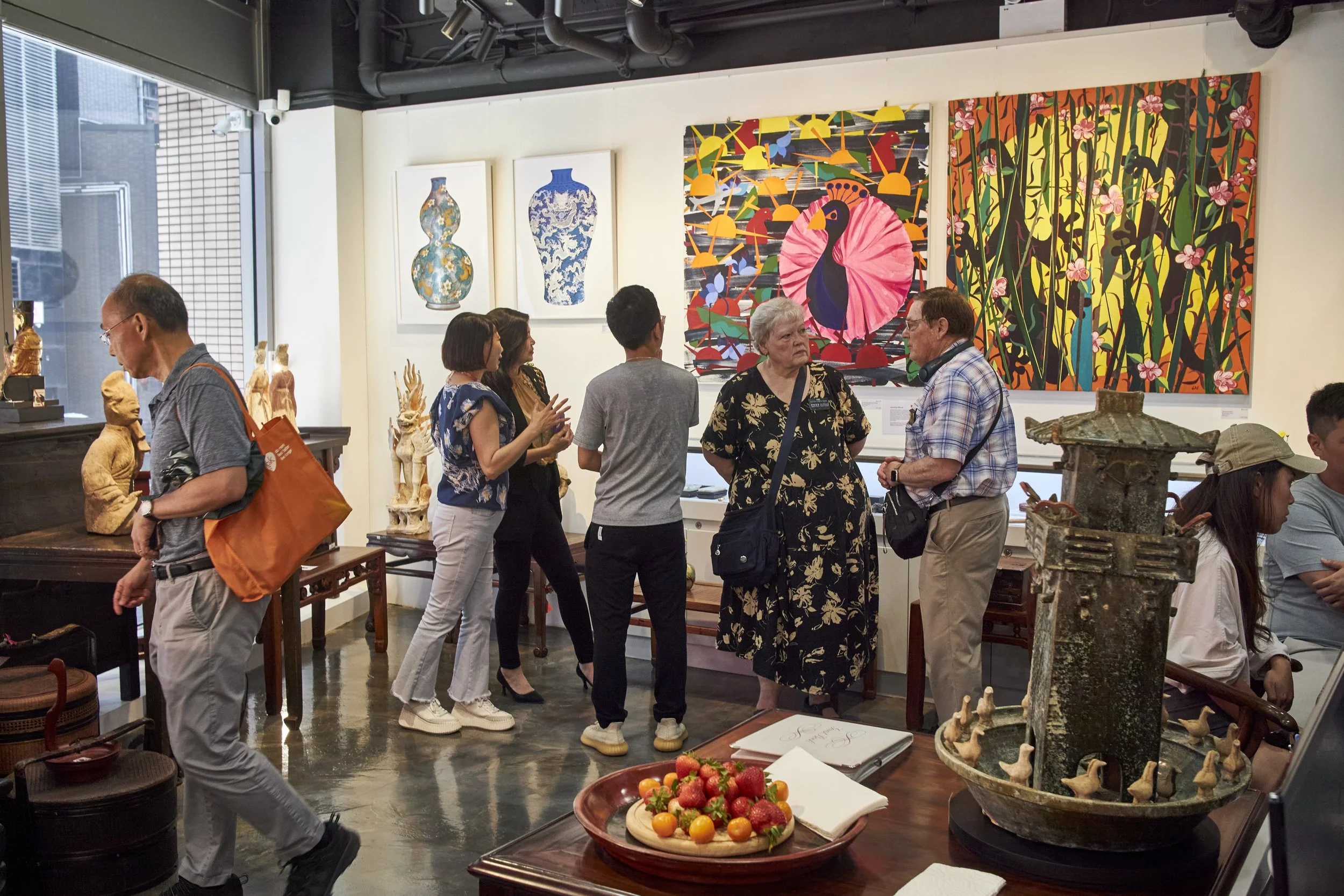 People viewing and discussing art in an art gallery, with paintings of vases, colorful abstract art, and sculptures on display.