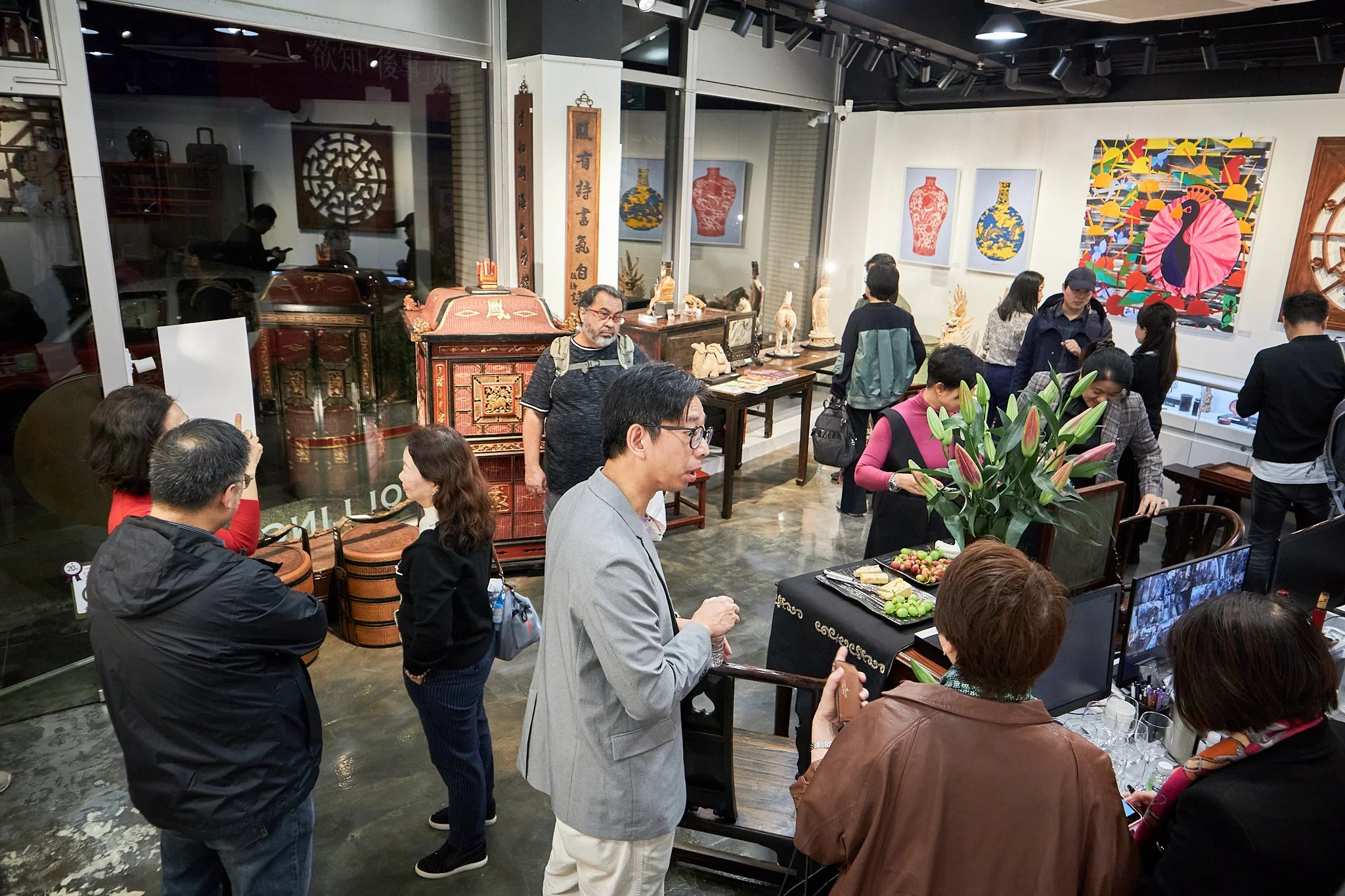 People browsing and interacting at an art gallery with Asian art, paintings of vases, sculptures, and traditional artifacts displayed on tables and walls.
