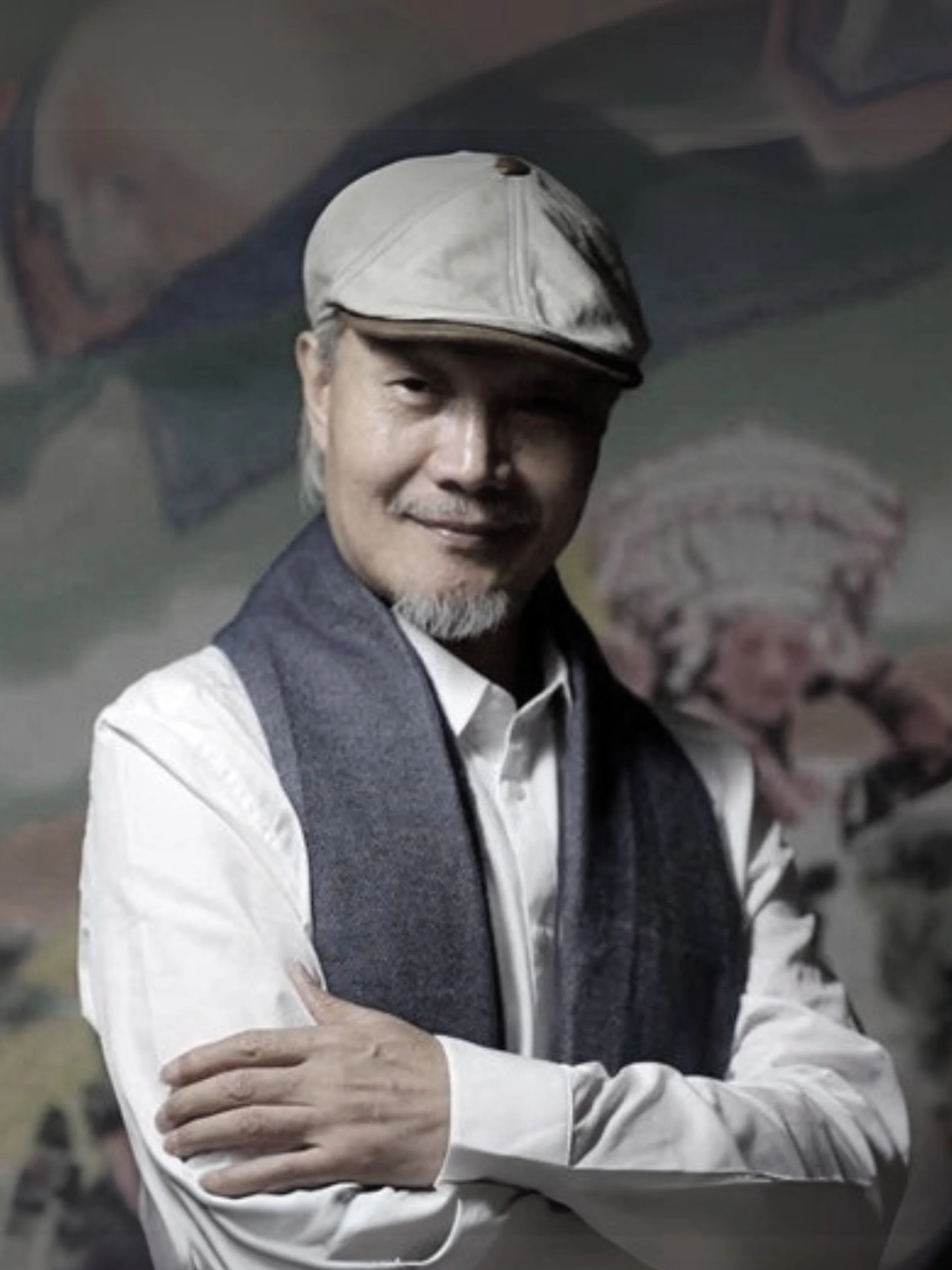 Fan Hongbin 樊鴻賓： A man with gray hair and beard, wearing a white cap, a white shirt, and a blue scarf, smiling with arms crossed in front of a colorful painted mural background.
