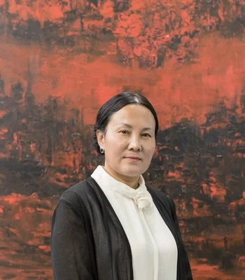 Zhou Lianhua 周連華：A woman with black hair and earrings, wearing a black blazer over a white high-neck blouse, standing in front of an abstract red and black background.