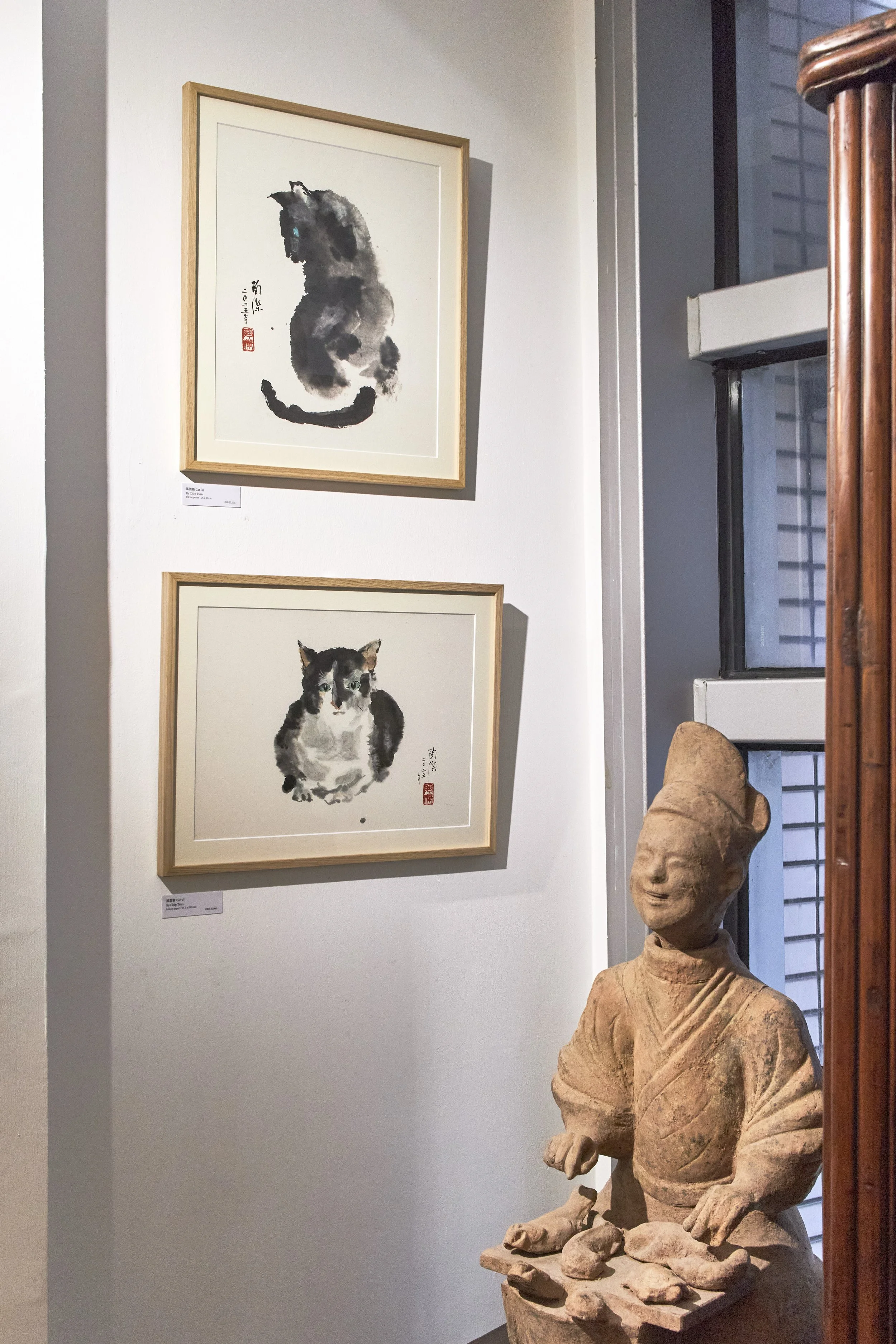 Gallery wall showcasing two framed watercolor paintings of cats, one sitting and one lying down, with Chinese characters and red stamps, alongside a stone sculpture of a smiling figure.
