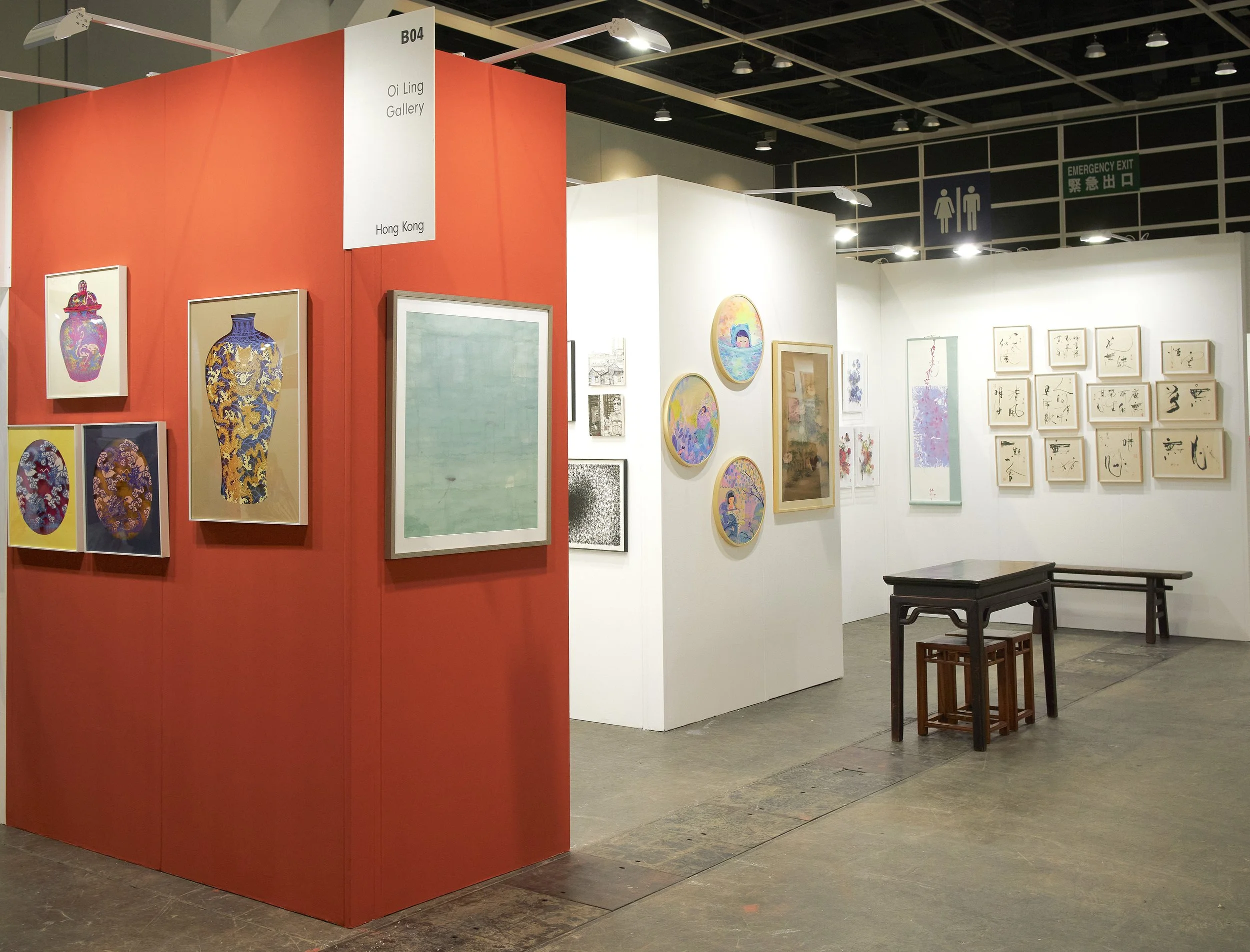 Affordable Art Fair 2025: featuring various paintings and artworks, including vases, circular landscape scenes, calligraphy, and mixed media pieces displayed on white and red walls, with a small table and stools in the foreground.