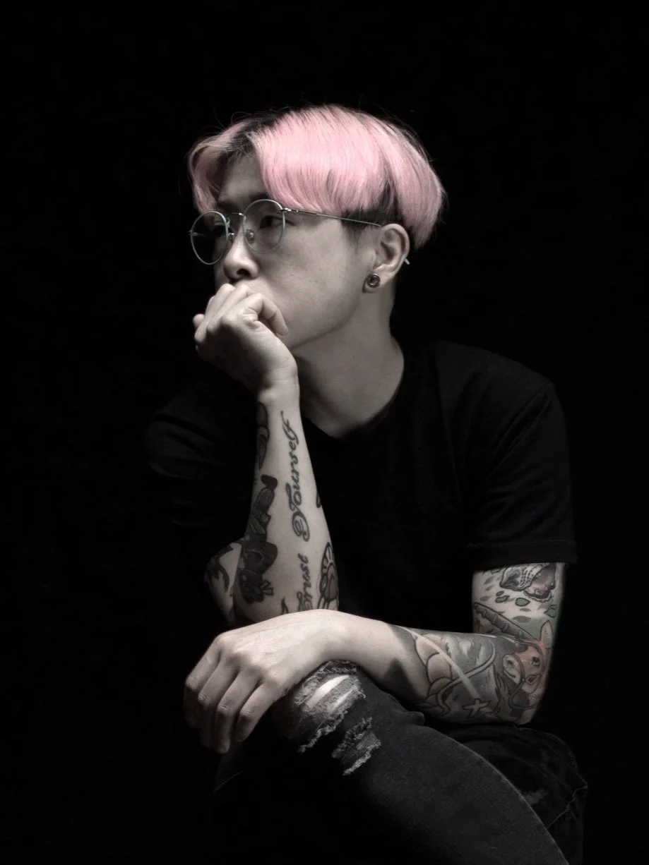 Zoie Lam 林雅儀: A young person with pink hair, glasses, and multiple tattoos on their arms, sitting against a black background, resting their chin on their hand in a thoughtful pose.