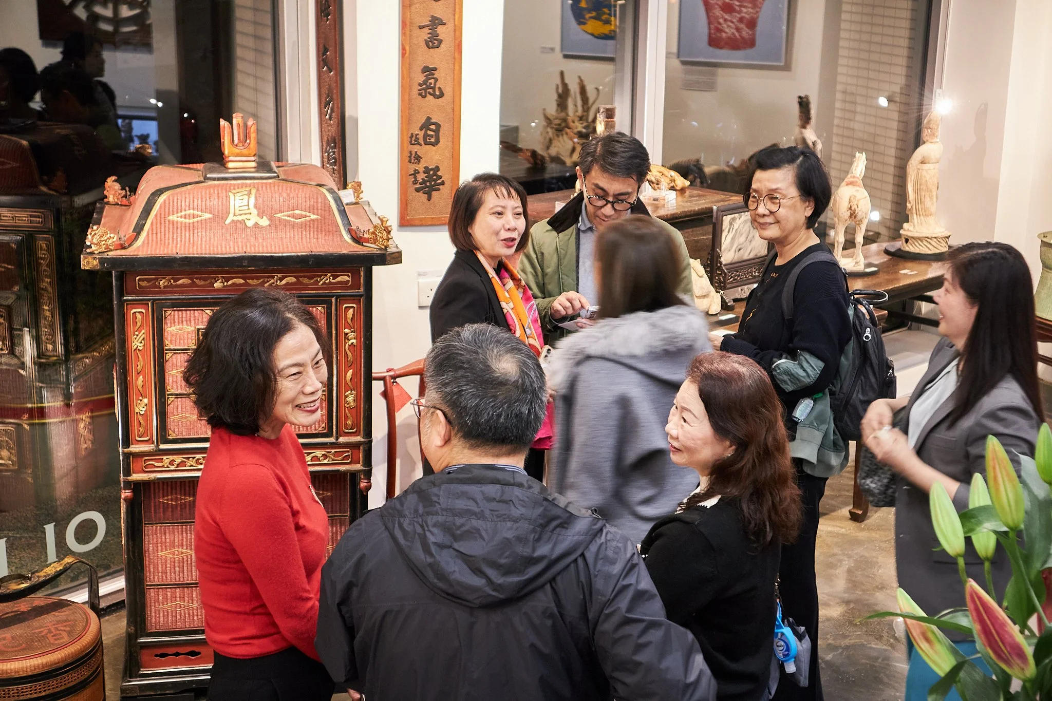Group of people socializing in an indoor art gallery or museum, surrounded by Asian art and artifacts, some of which are displayed on shelves and walls.