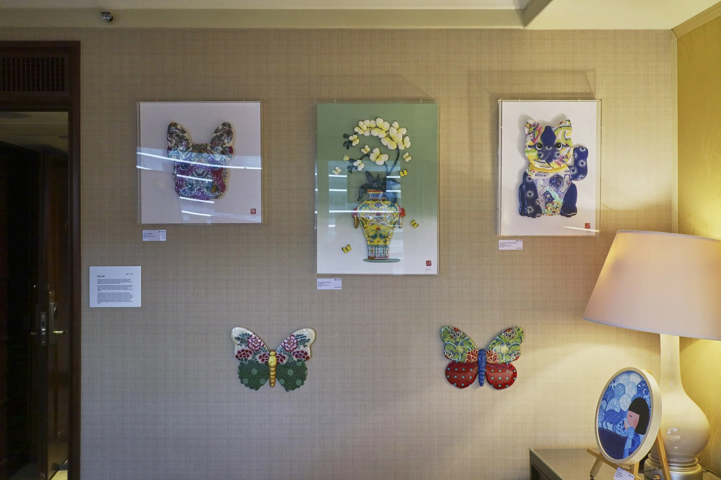 An art display on a beige wall featuring three framed artworks and two butterfly-shaped fabric art pieces. The artworks include a colorful butterfly face, a vase with flowers, and a patterned dog face. There is a table with a lamp and an oval frame w