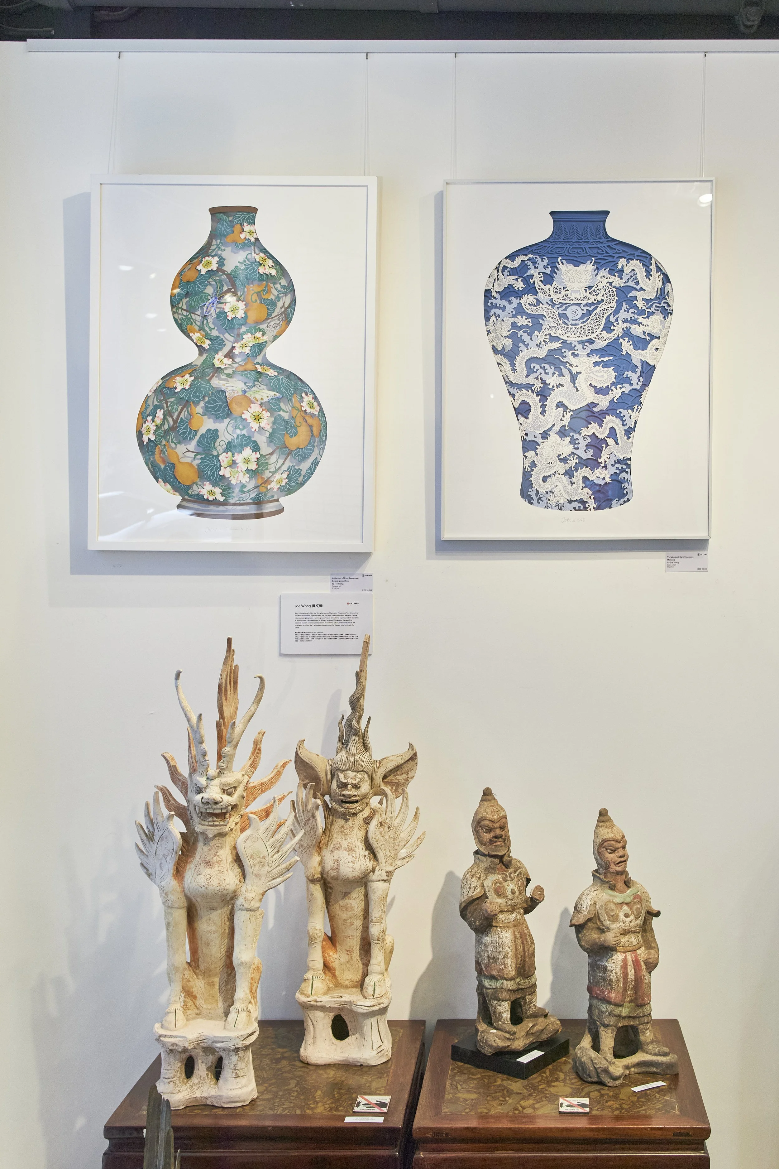 Two framed paintings of vases hang on a white gallery wall, with colorful floral and dragon designs, and a pair of carved wooden statues resembling mythical creatures and warriors displayed on separate tables below.