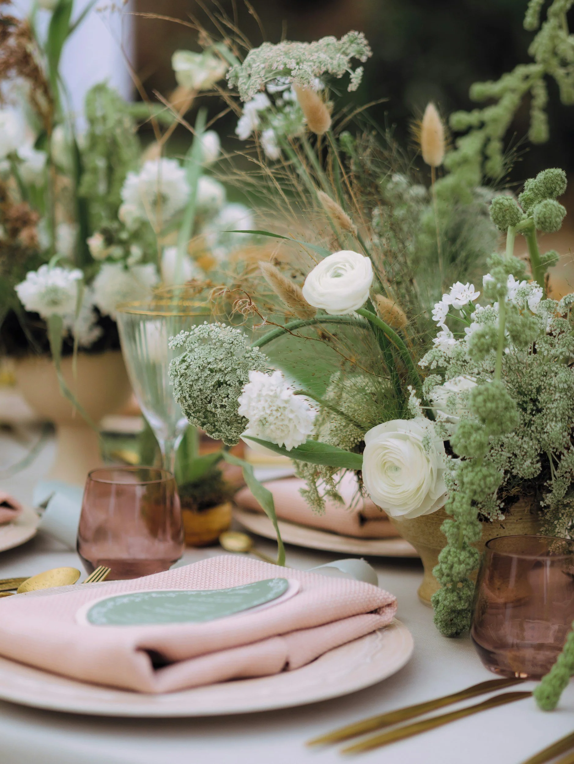 A close-up of an elegant table setting with a pink napkin, a plate, and two pink-tinted glasses, decorated with a lush floral centerpiece featuring white roses, green foliage, and dried grasses.