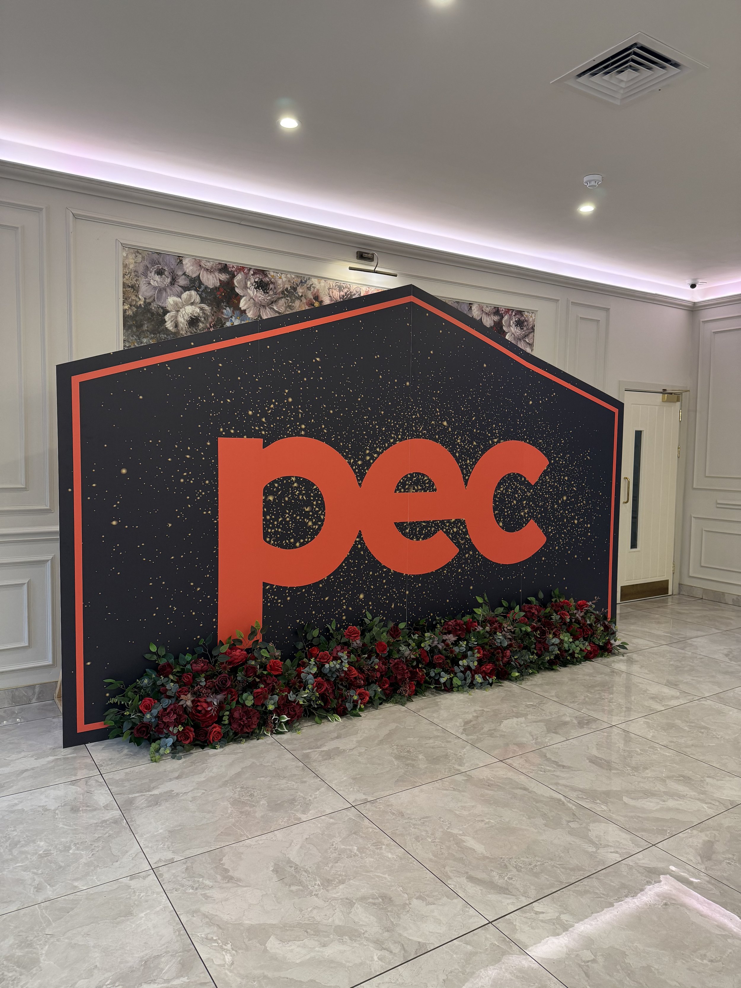 A black display board with the word 'pec' in large red letters, decorated with a red border, and surrounded by a bed of red and green flowers at its base. The background features a floral tapestry and an interior wall with white paneling, in a well-lit room.