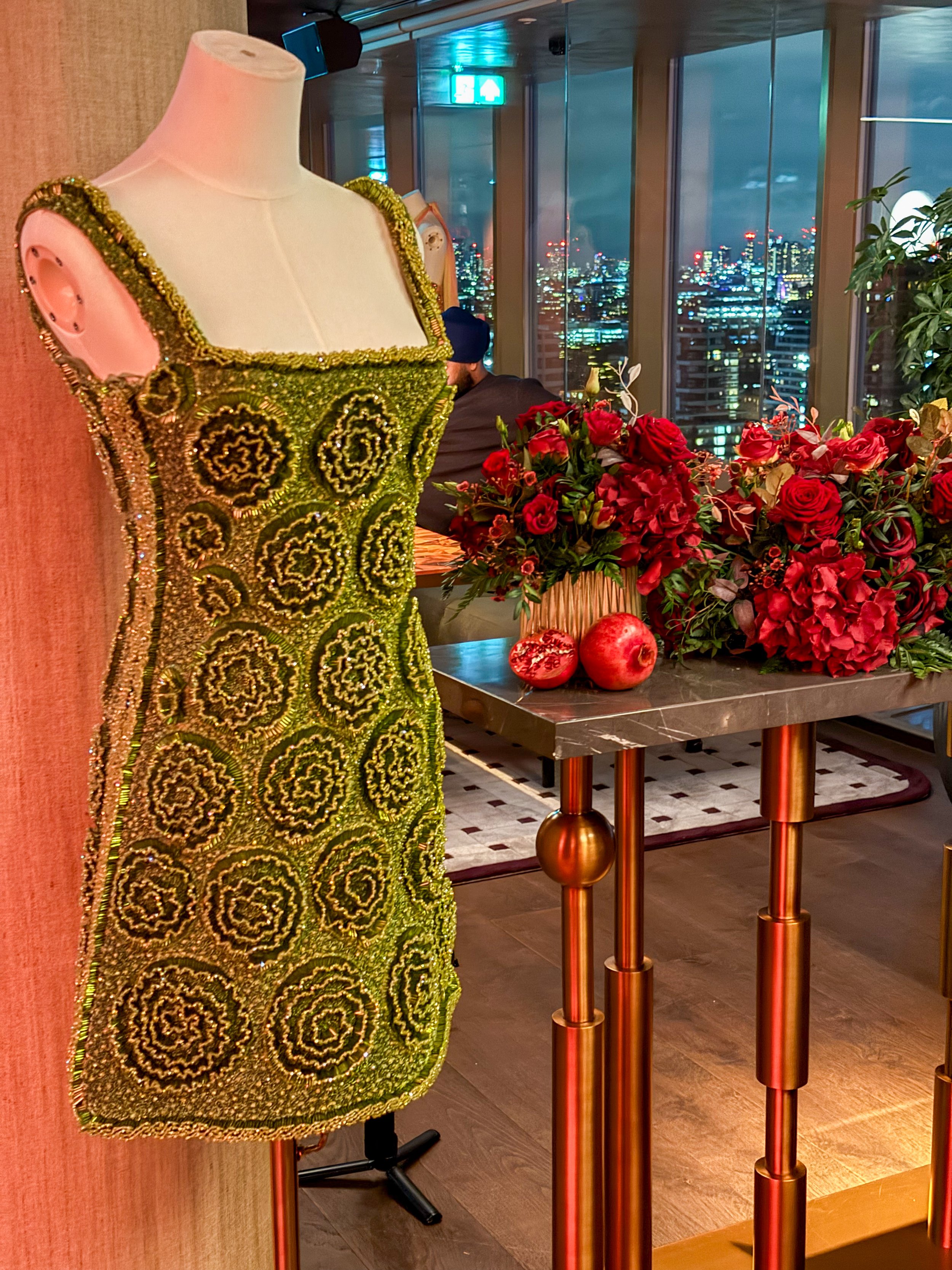 A mannequin wearing a green and gold beaded dress in a room with a city view through floor-to-ceiling windows, a table with red roses and pomegranates, and a person sitting in the background.