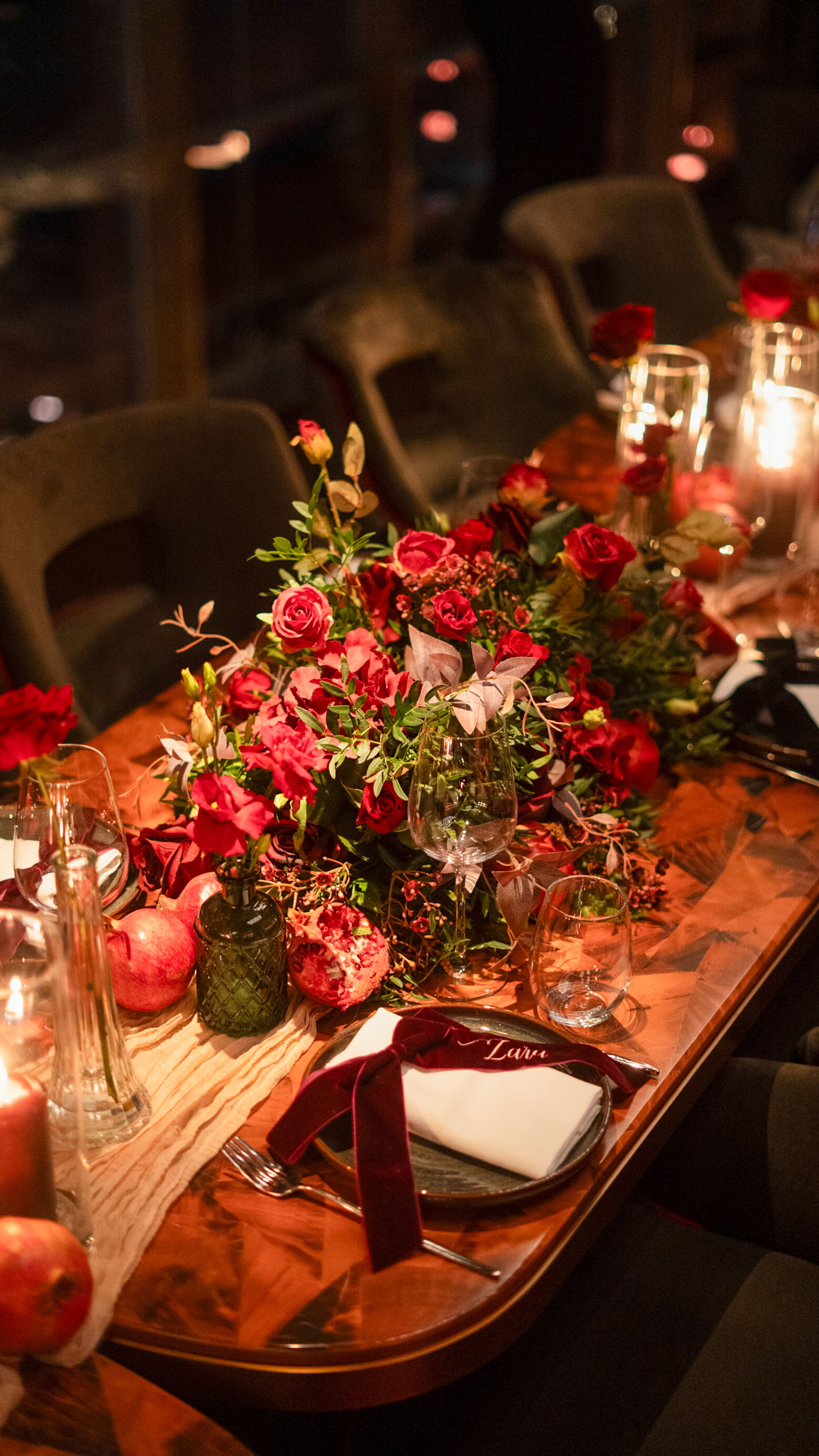 A decorated dining table with a floral centerpiece of red and pink roses, candles, apples, and bottles, set for a meal in a dimly lit room.