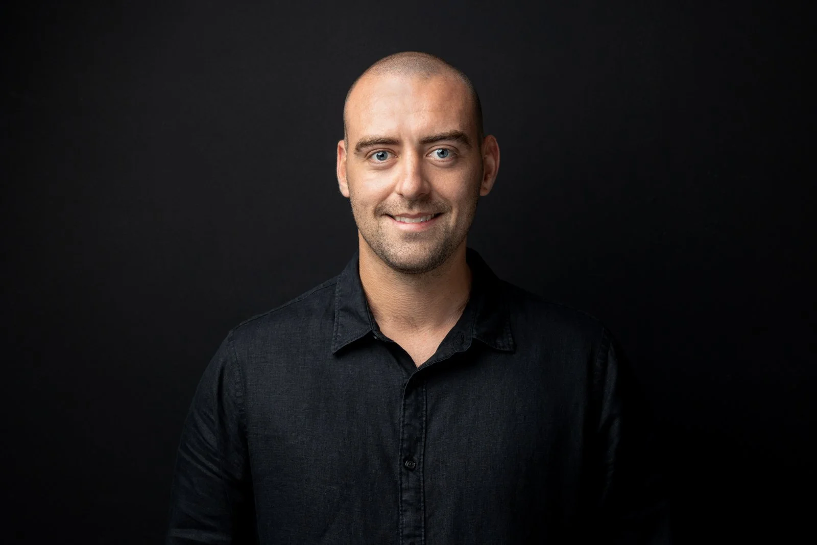 Headshot of a smiling man with a shaved head wearing a black shirt against a dark background.