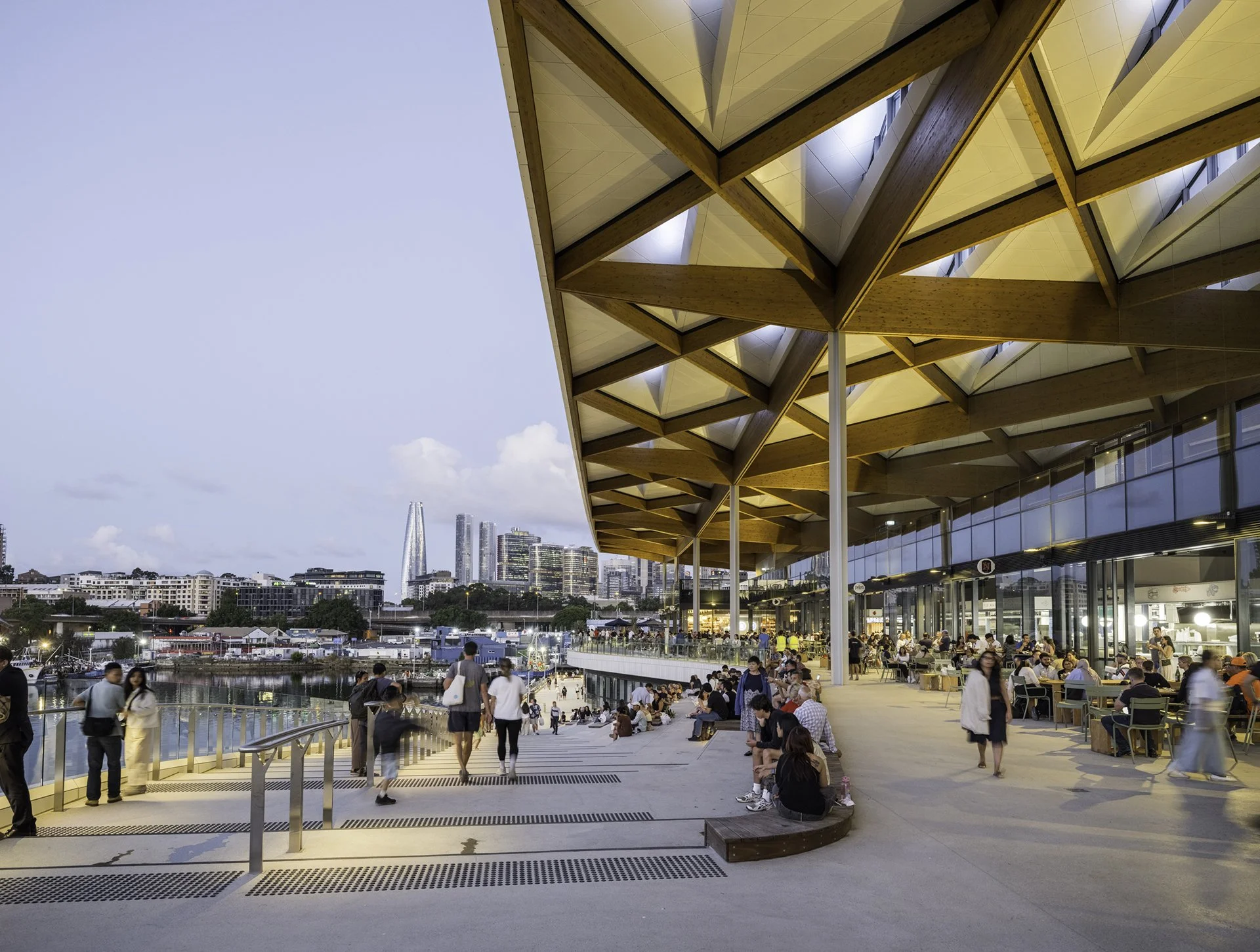 People walking and sitting outside a modern building with a large overhang, overlooking a waterfront with boats and a city skyline in the background during evening.