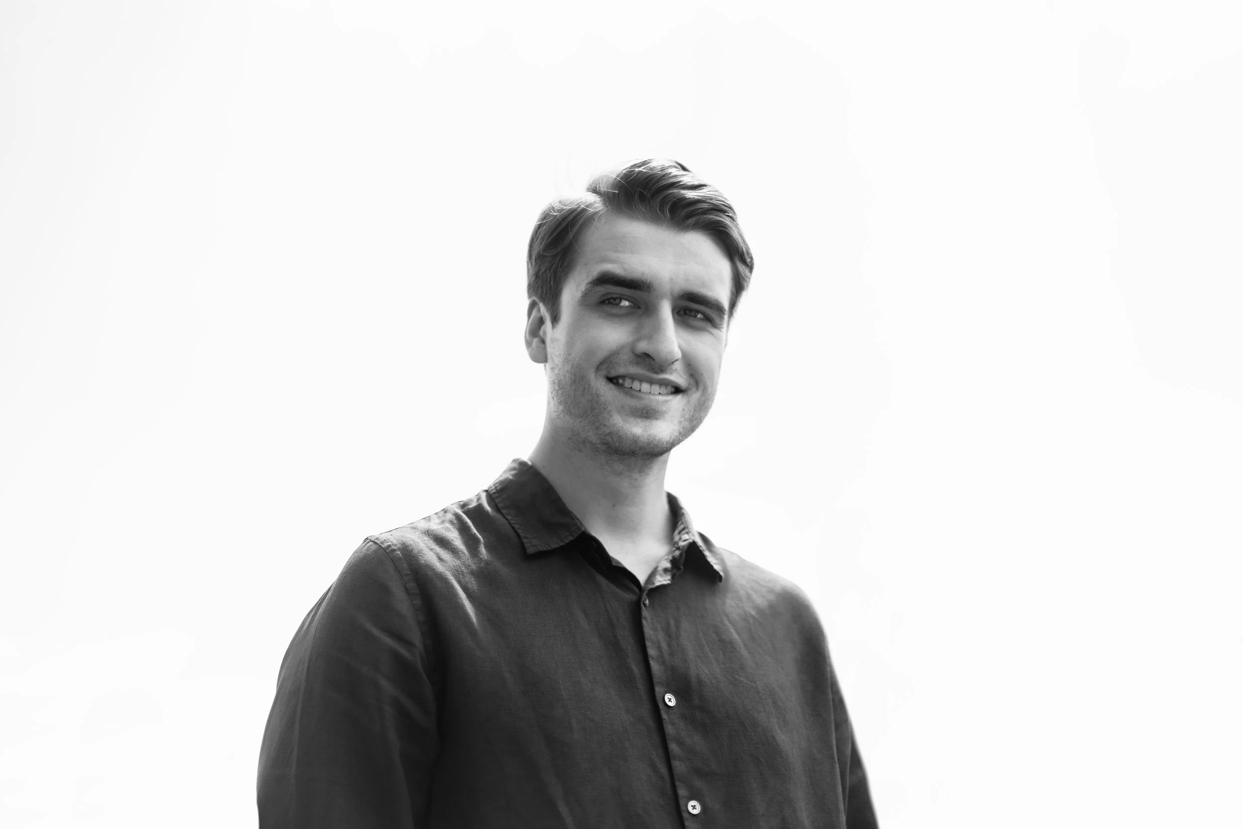 Black and white portrait of a young man with styled hair, wearing a dark button-up shirt, smiling softly against a plain light background.