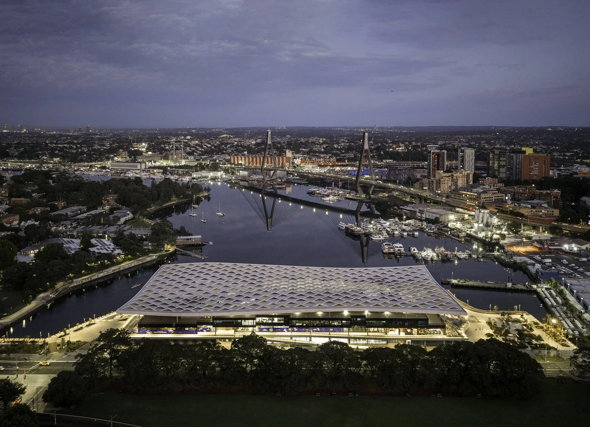 Nighttime cityscape featuring a unique cable-stayed bridge over a river, with high-rise buildings in the background and a large stadium or convention center with a patterned roof in the foreground.