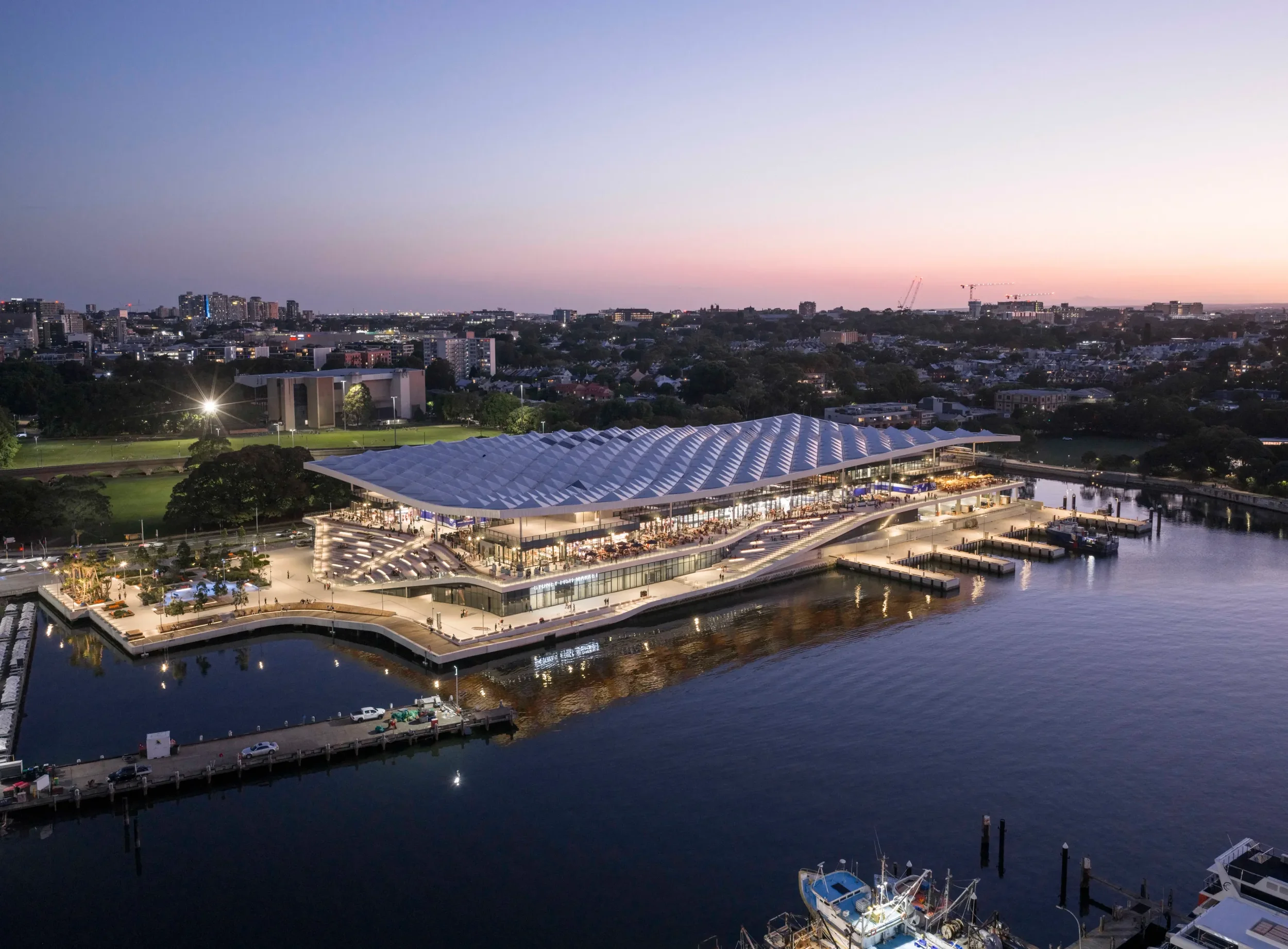 A cityscape at sunset featuring a large modern building by a waterway with boats docked, surrounded by trees and high-rise buildings in the distance.