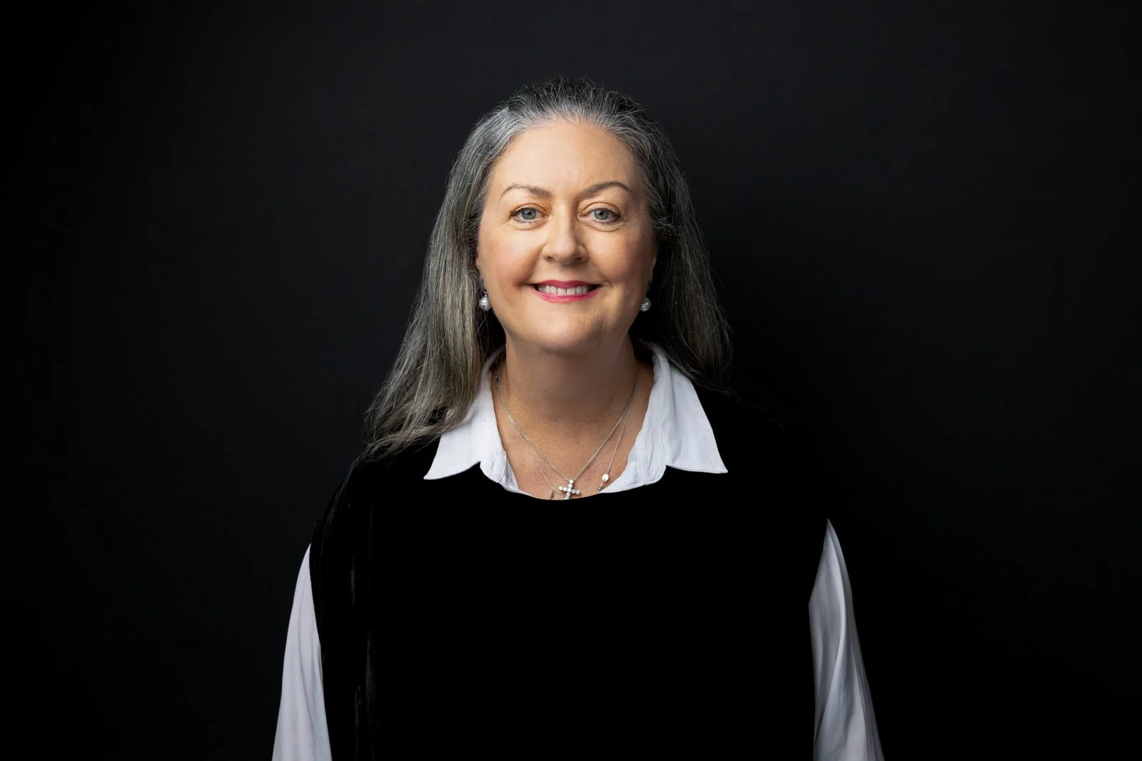 A middle-aged woman with long gray hair, wearing a white collared shirt under a black sweater, and pearl earrings, smiles against a black background.