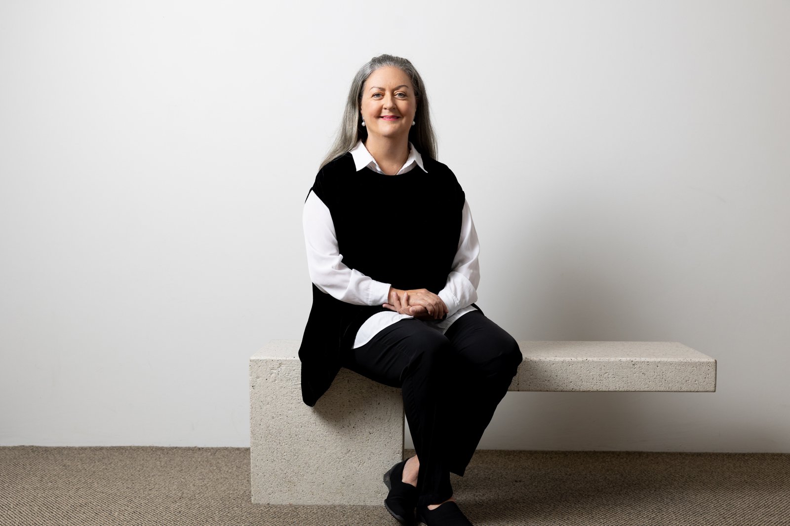 A woman with gray hair sitting on a concrete block against a plain white wall, smiling and looking at the camera, wearing a white blouse and black vest.