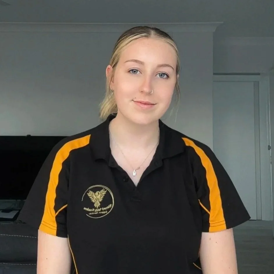 A young woman with blonde hair tied back, wearing a black polo shirt with yellow accents and an embroidered phoenix, sitting in a room with light-colored walls and a door in the background.