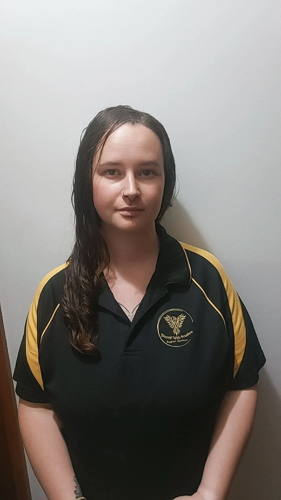 A young woman with long wet dark hair, pierced nose and lip, wearing a black polo shirt with gold accents, standing against a plain white wall.