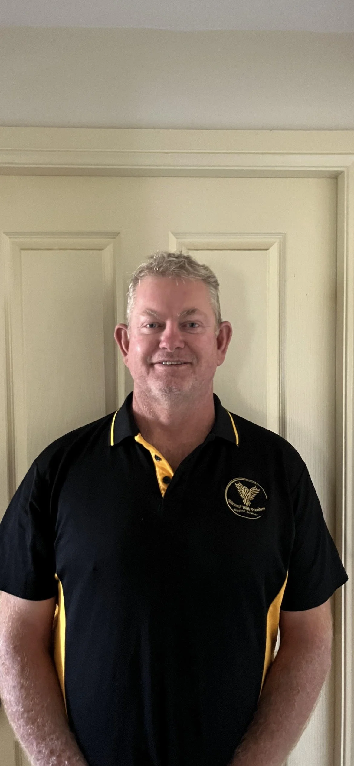 A man with short, curly gray hair standing in front of a beige door, wearing a black sports polo shirt with yellow accents and a logo.