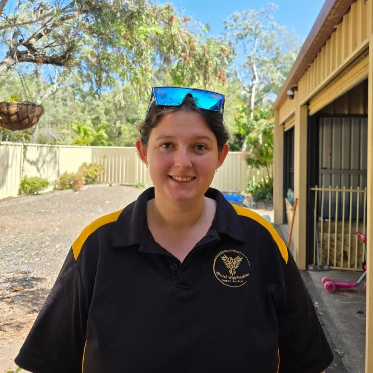 A young woman with short dark hair and light skin smiling outdoors on a sunny day, wearing sunglasses on her head and a black polo shirt with yellow accents and a logo on the left side.