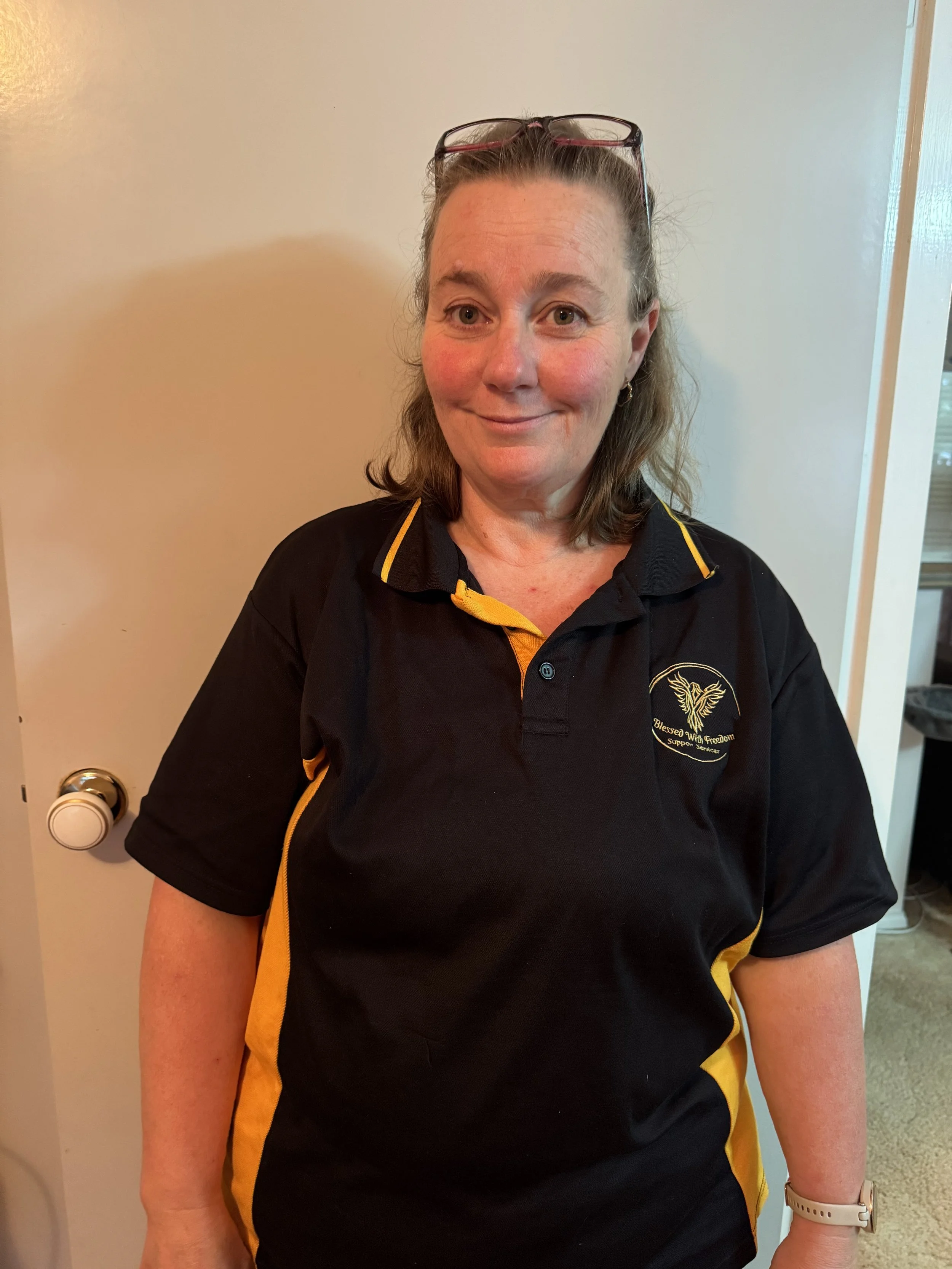 A woman with shoulder-length hair, wearing glasses on her head and a black and yellow sports shirt, standing indoors near a door.