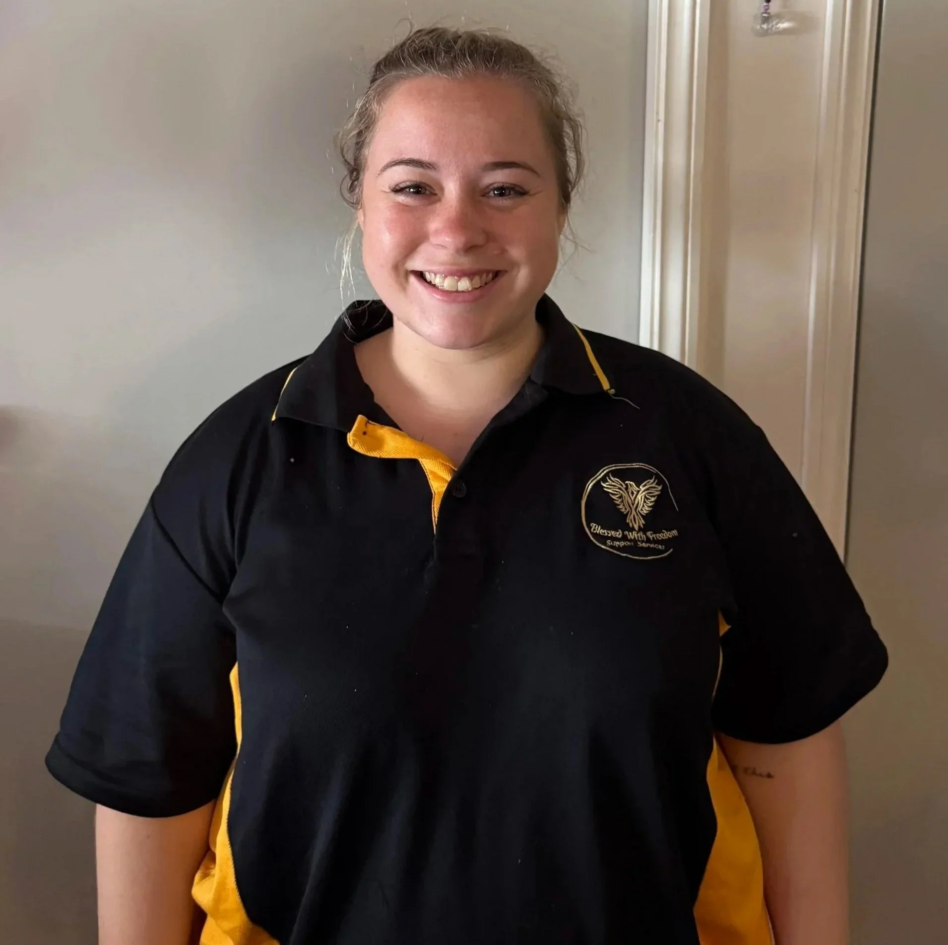 A young woman with blonde hair smiling, wearing a black and yellow uniform polo shirt with an embroidered logo featuring a butterfly and the text "Blessed With Freedom Support Services"