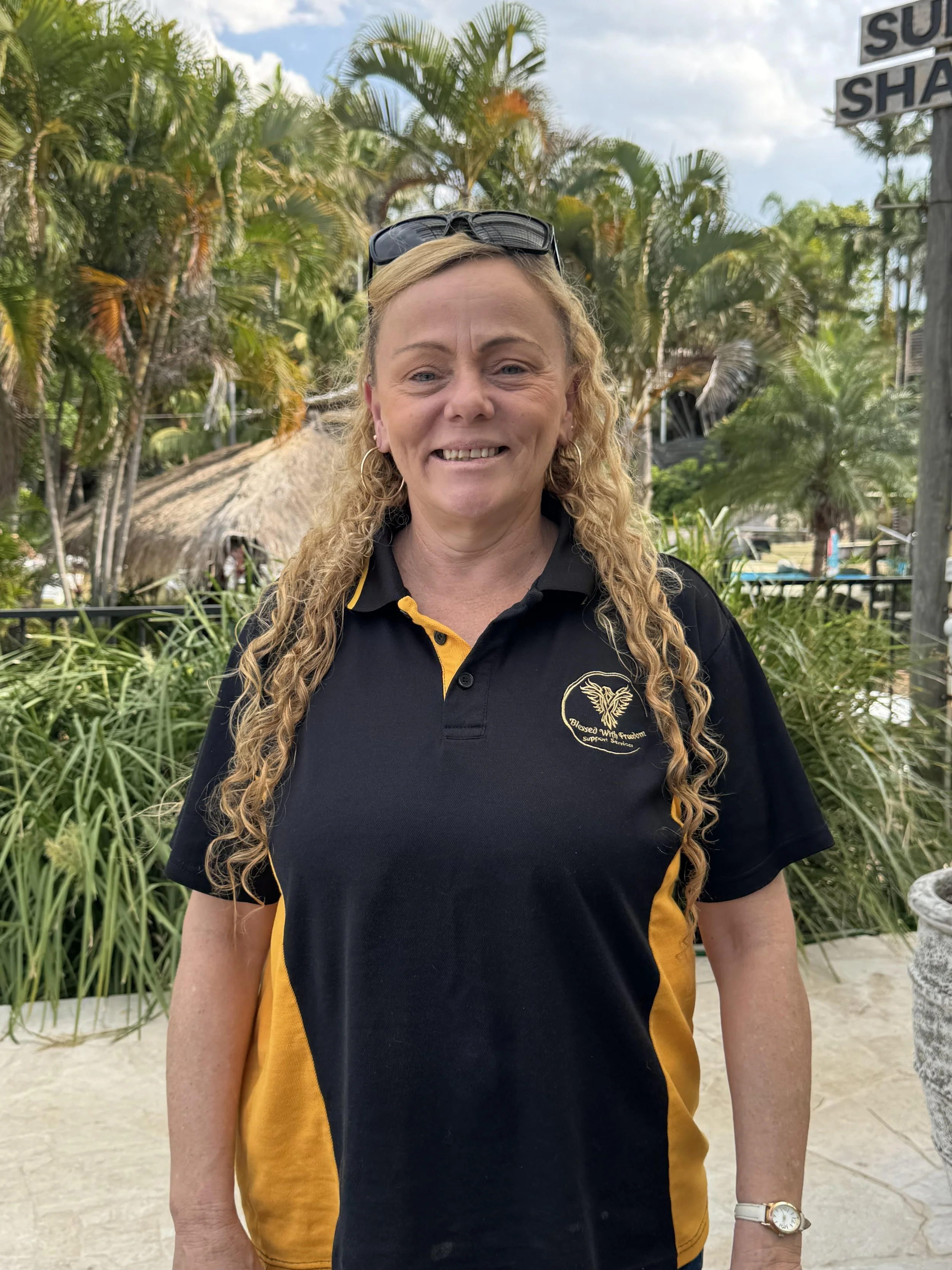 A woman with curly blonde hair, wearing sunglasses on her head, a black and yellow polo shirt, standing outdoors with tropical plants and palm trees in the background.