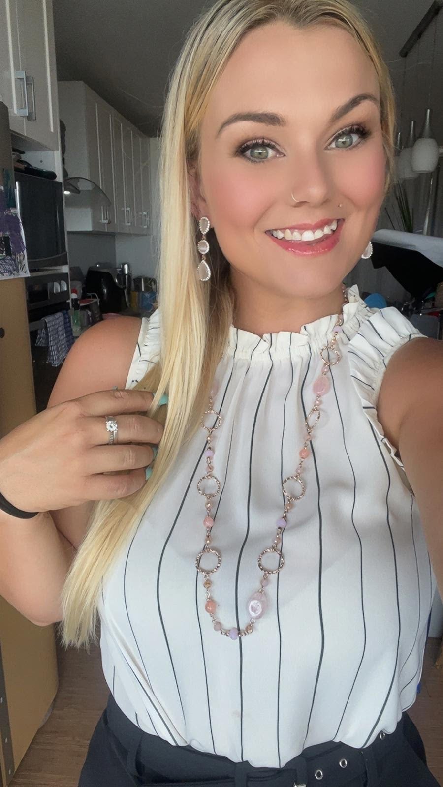 A smiling woman with long blonde hair, wearing a business attire, white sleeveless top with black vertical stripes, and jewelry including earrings, a necklace, a ring, and a bracelet.