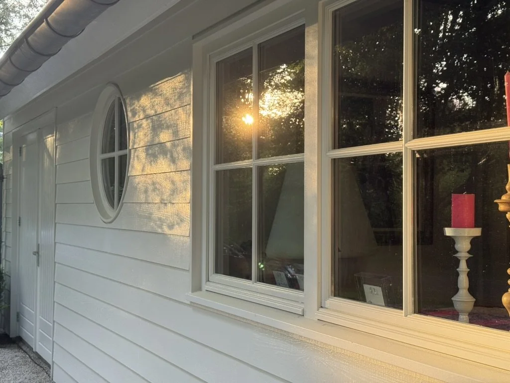 Outside of a white house with a circular window and a larger rectangular window, sunlight shining through the trees