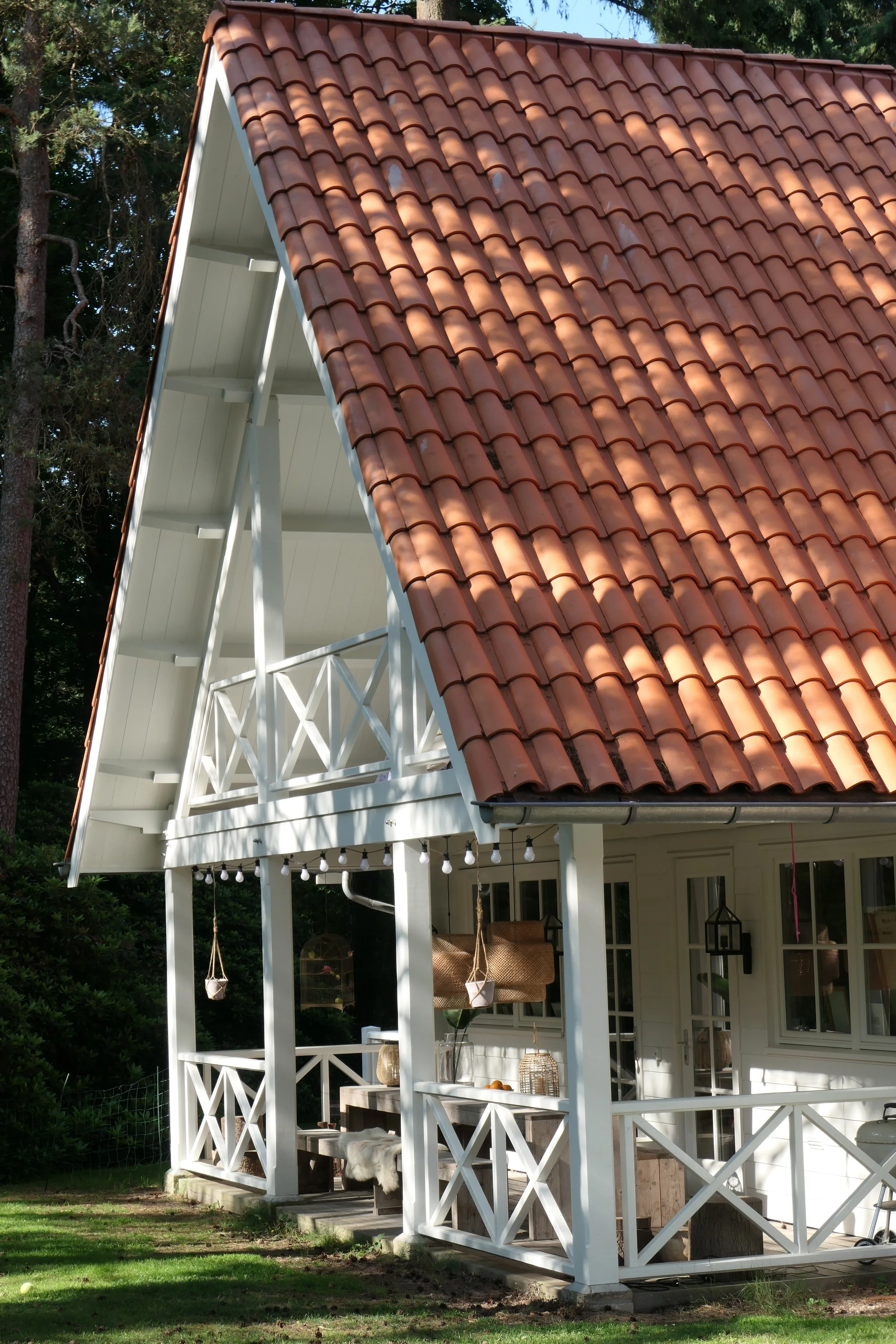 A house with a red tiled sloped roof, white wooden balcony and porch, surrounded by trees and greenery.