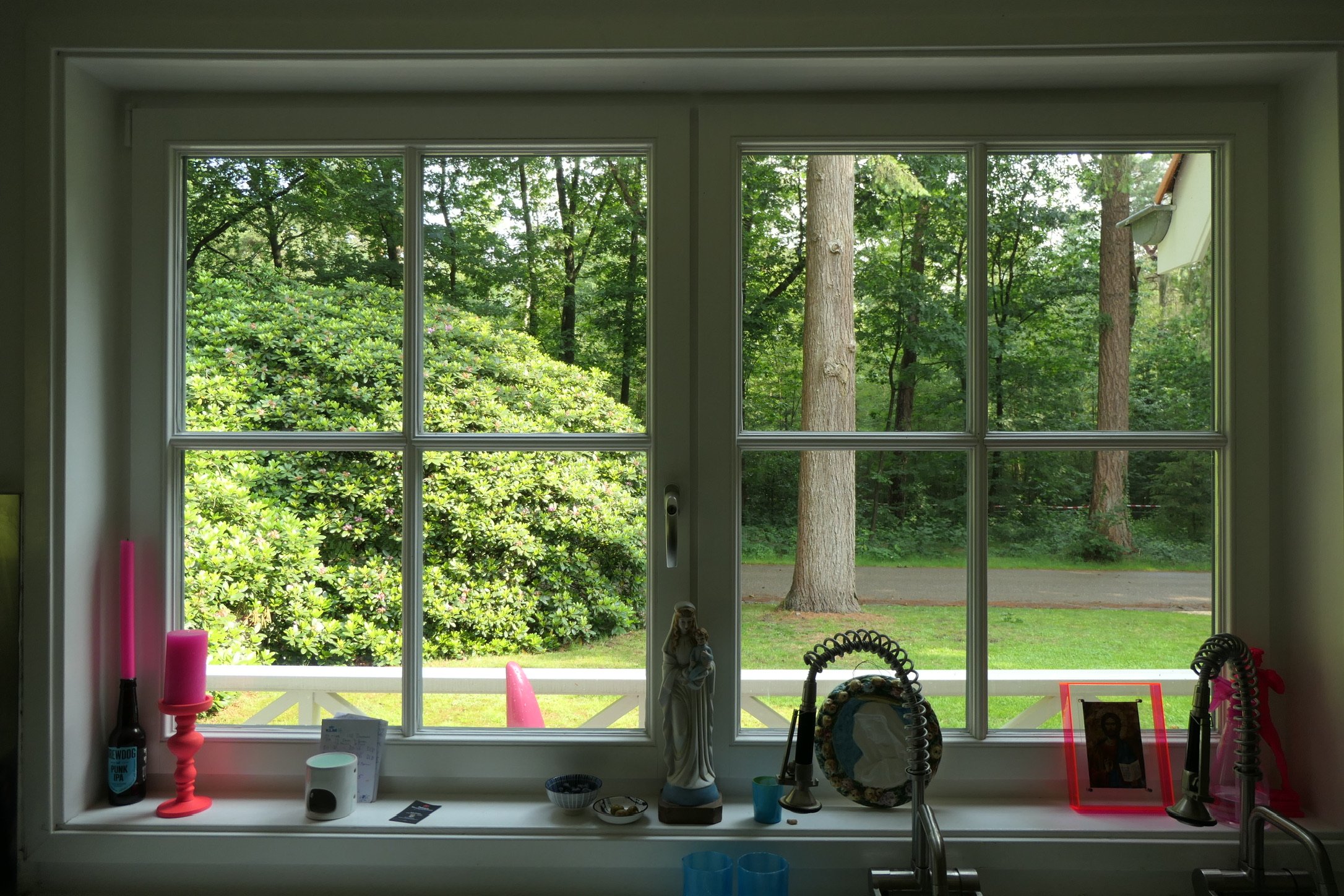 View through a kitchen window showing a lush green yard with trees and shrubs outside. The windowsill has various objects including pink candles, a small statue, a framed picture, bowls, and kitchen fixtures.