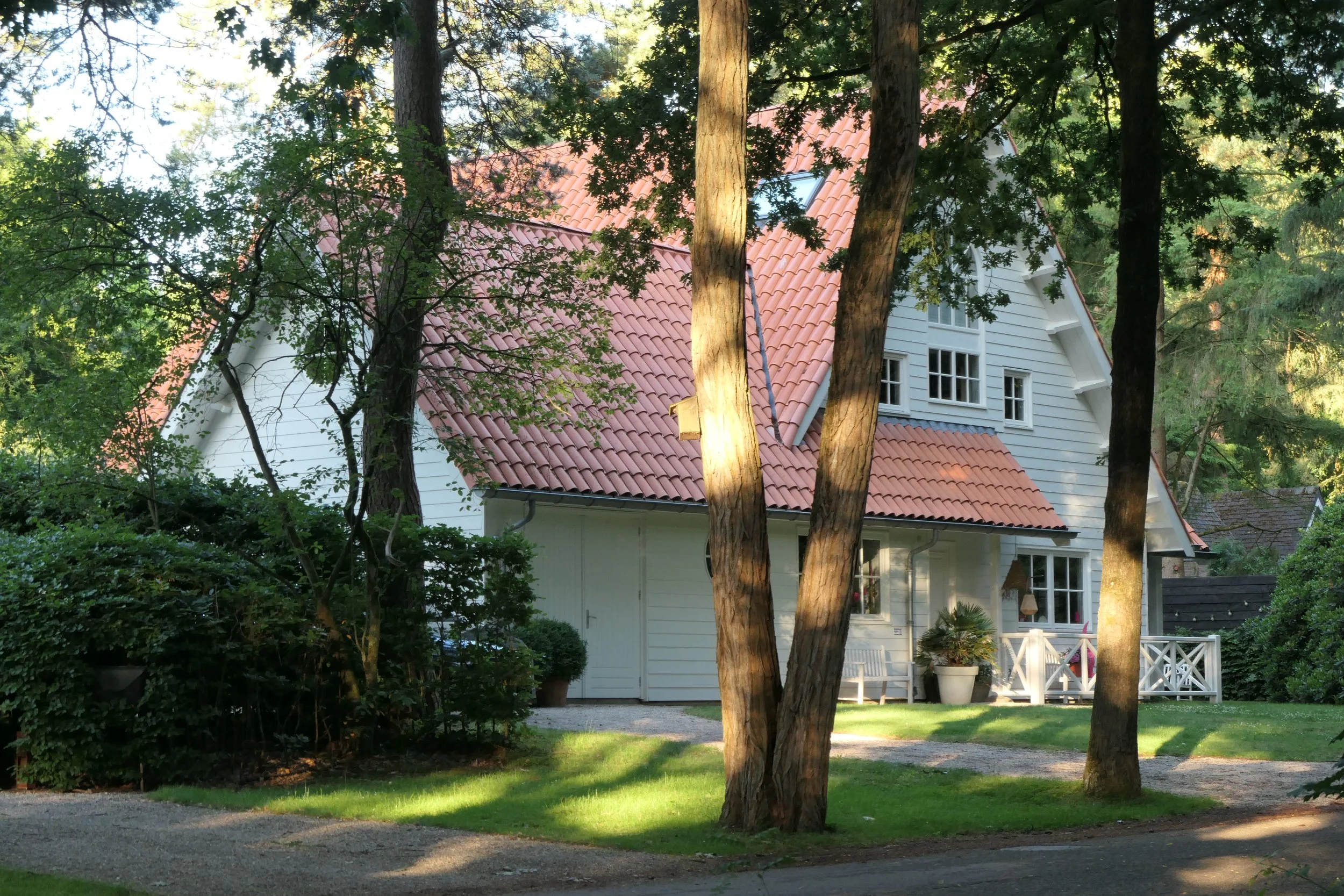 A white house with a red tile roof surrounded by trees and greenery, with a small front porch and potted plants.
