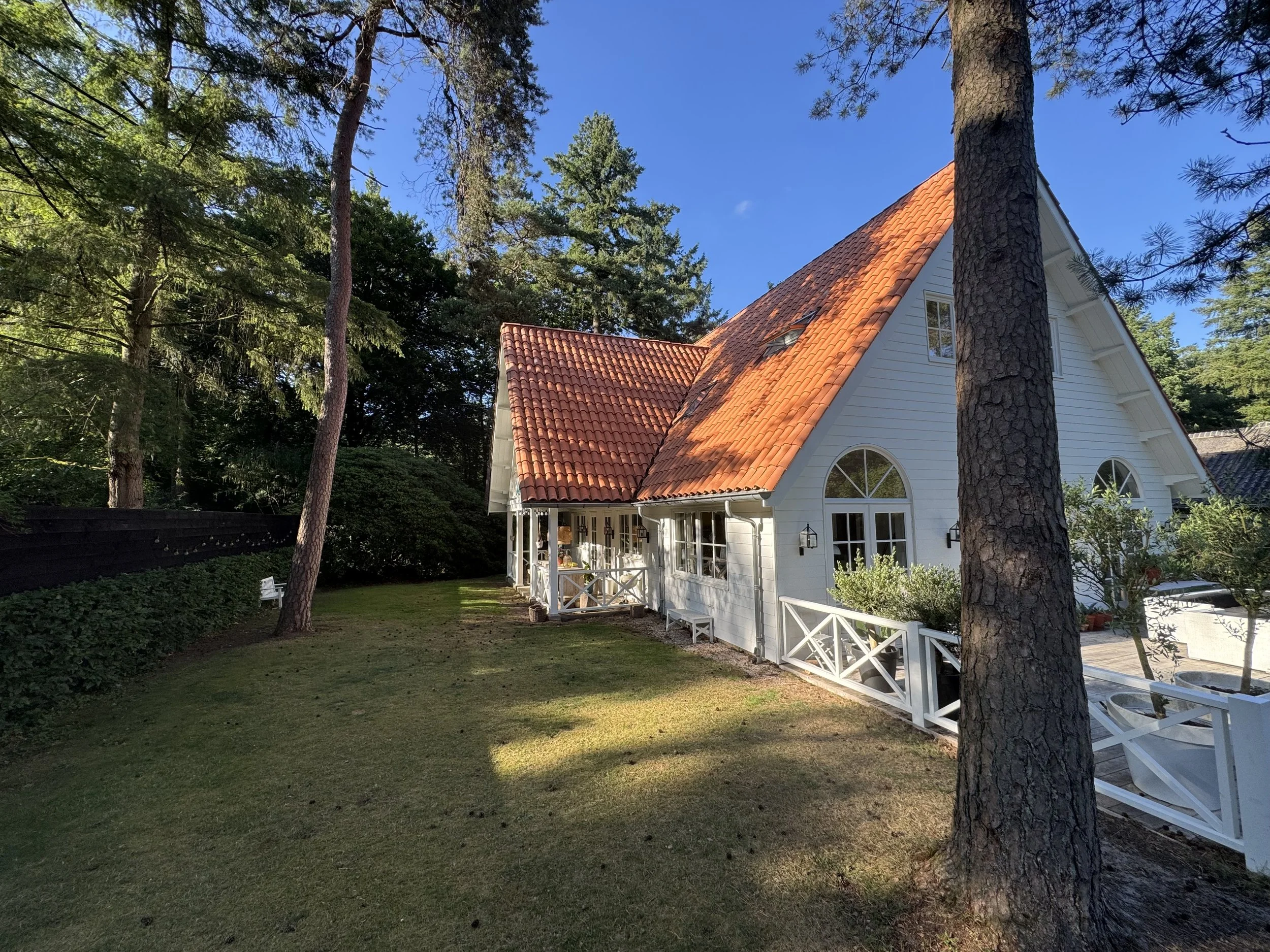 Charming white house with an orange tiled roof, surrounded by tall trees and a well-maintained lawn in a peaceful garden setting.