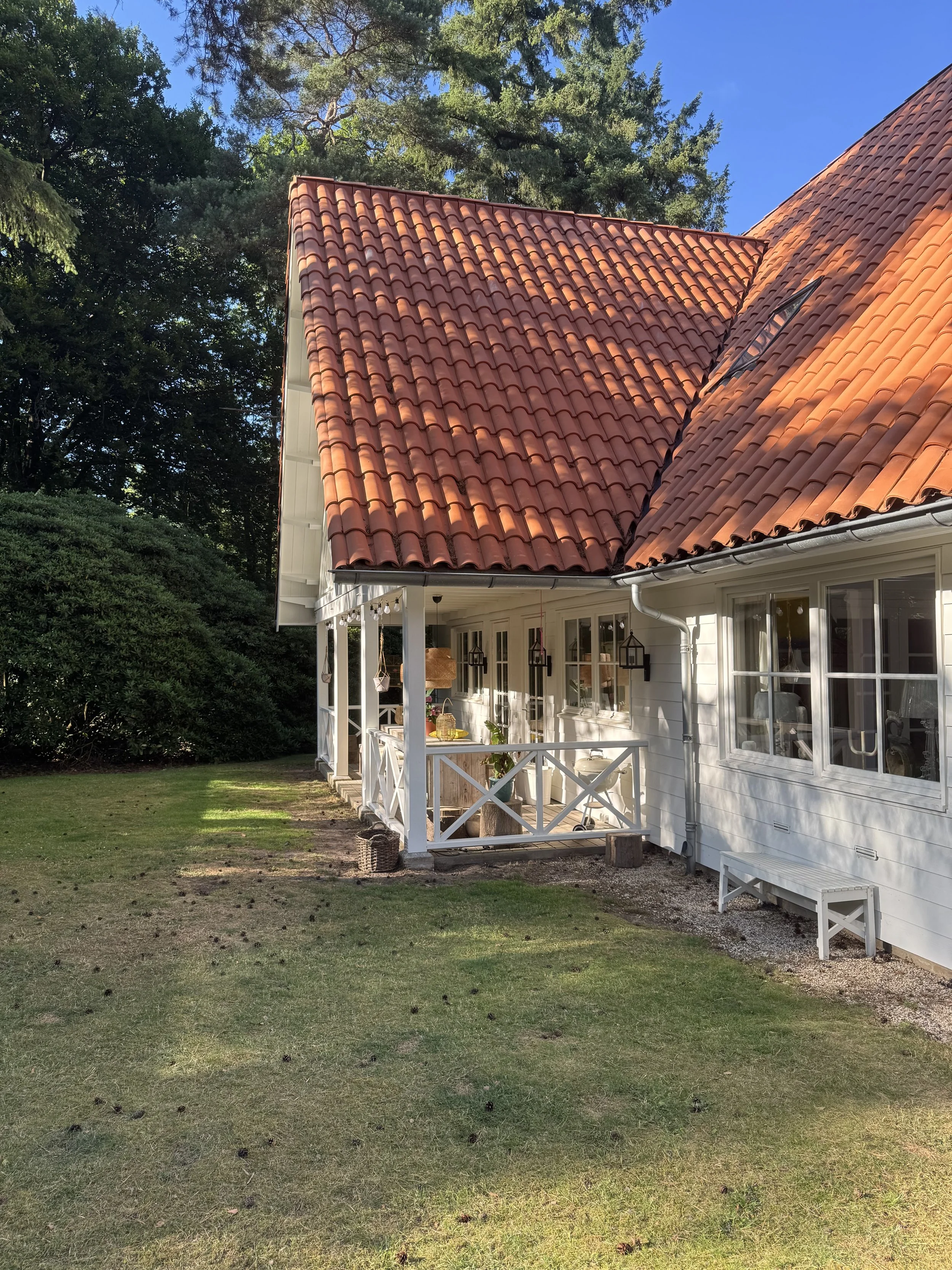 White house with a red tile roof, small porch with white railing, and green lawn with bushes and trees in the background.