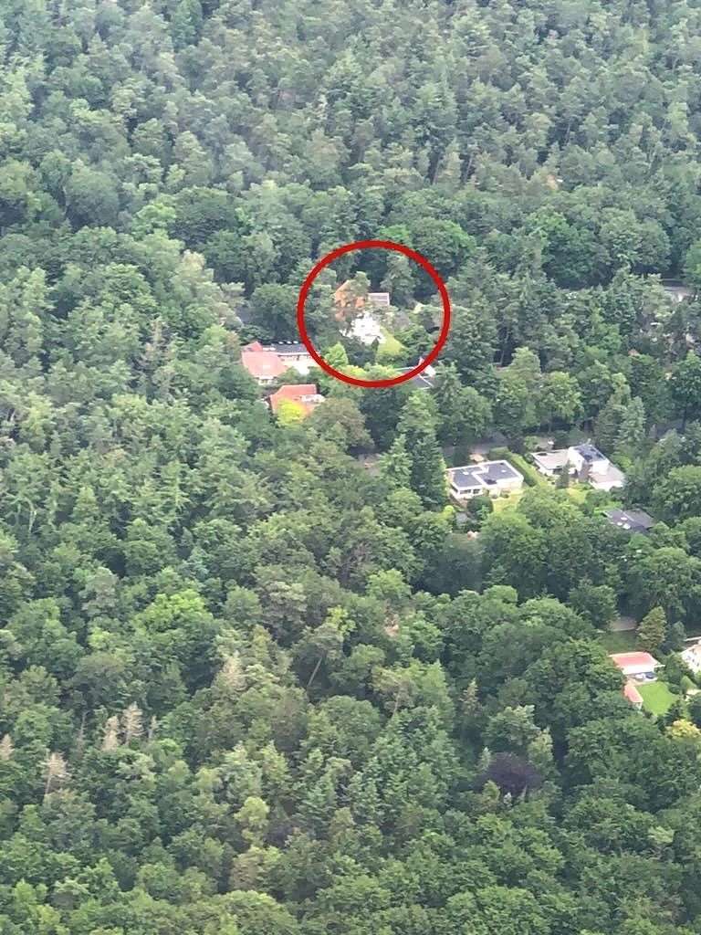 Aerial view of a heavily wooded area with a few houses visible among the trees, one house is outlined with a red circle.