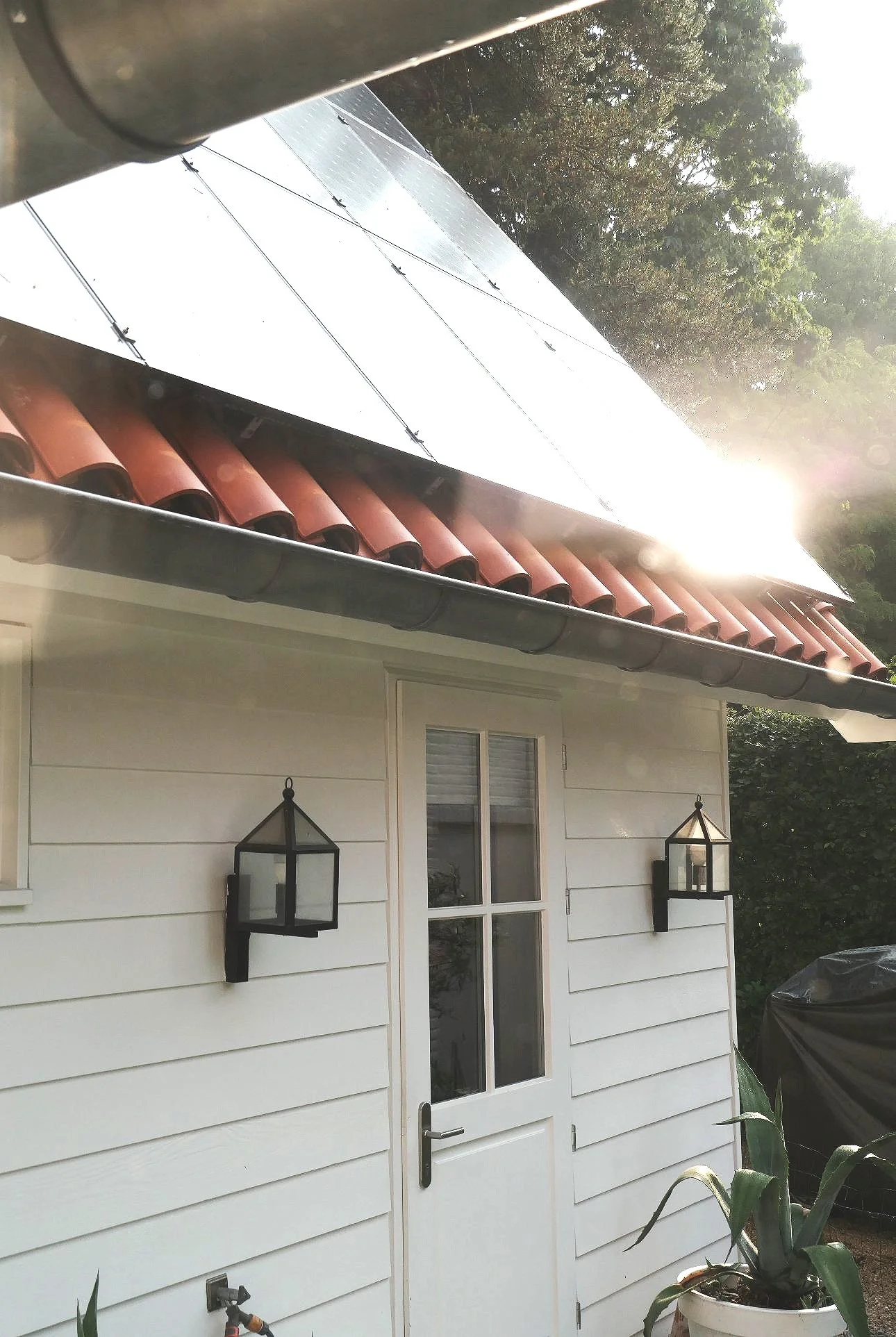 Exterior of a white house with a door, two black-framed lanterns, and a potted plant, with a solar panel on the roof and sunlight shining.