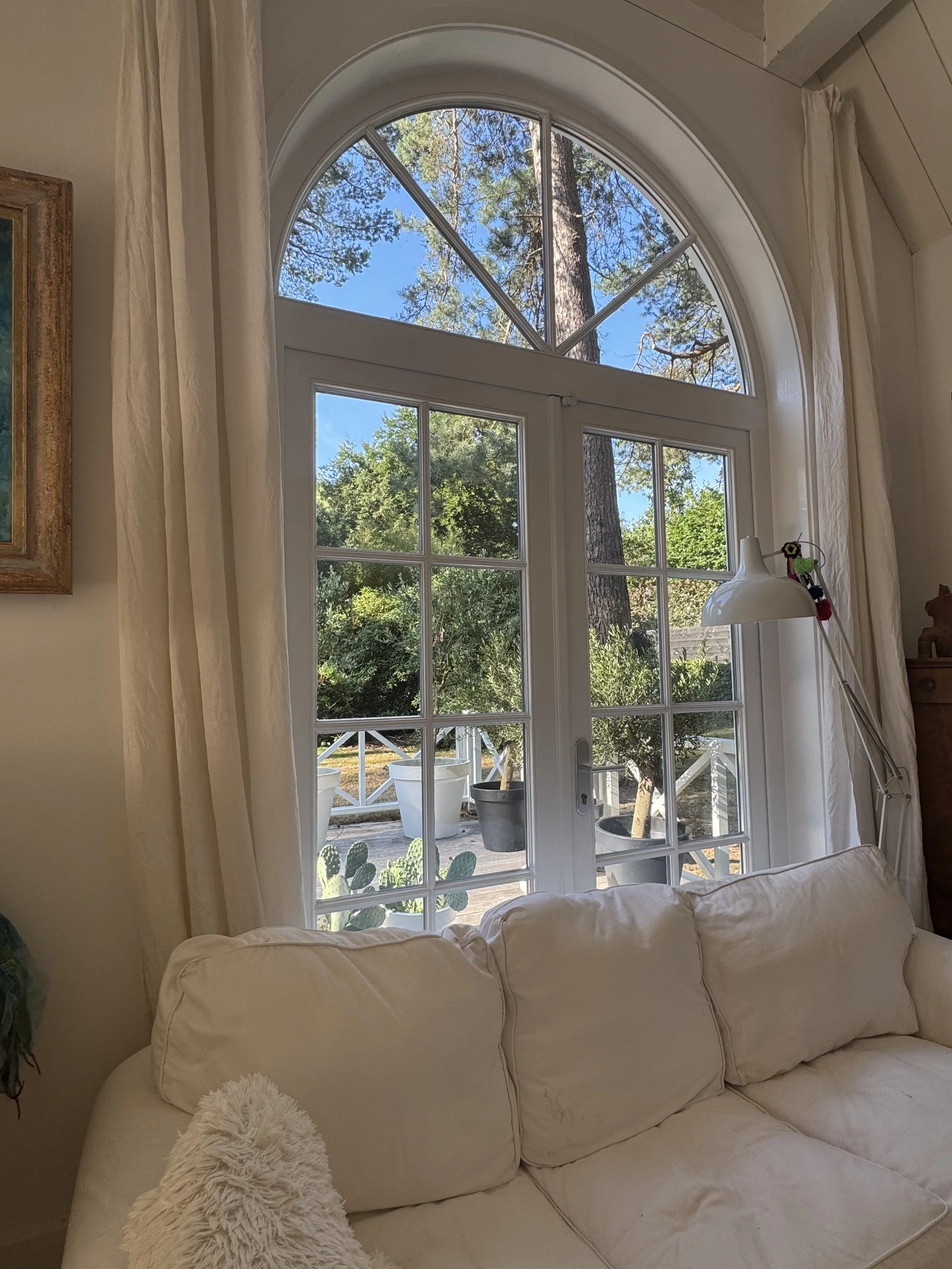 Living room with cream-colored sofa, large window with curtains, view of outdoor patio with potted plants, trees, and a blue sky.