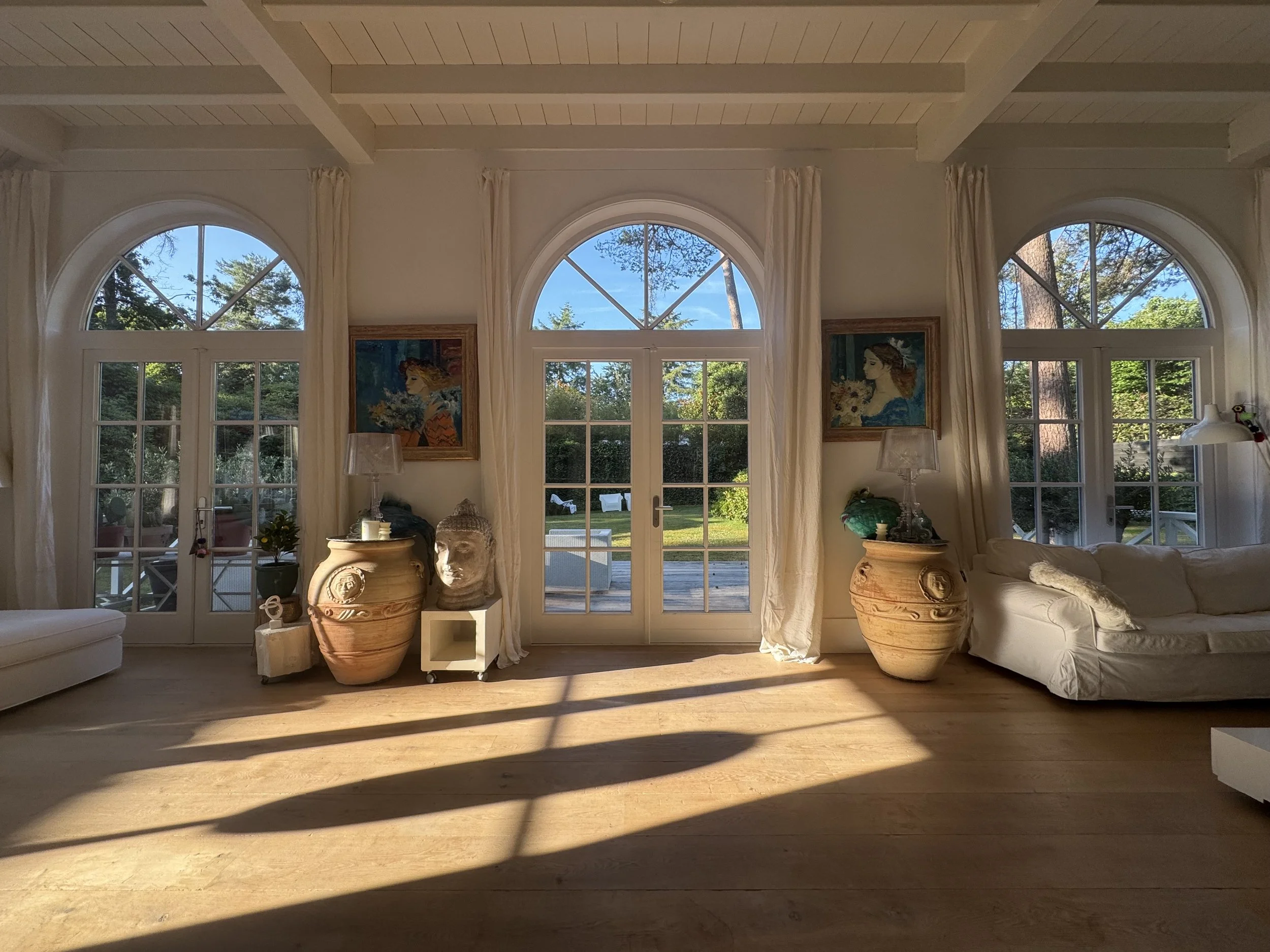 Living room with three large arched windows, white curtains, white walls, wooden floors, and furniture including a white sofa. There are decorative items such as large potted plants, a stone face sculpture, and framed artwork on the walls.