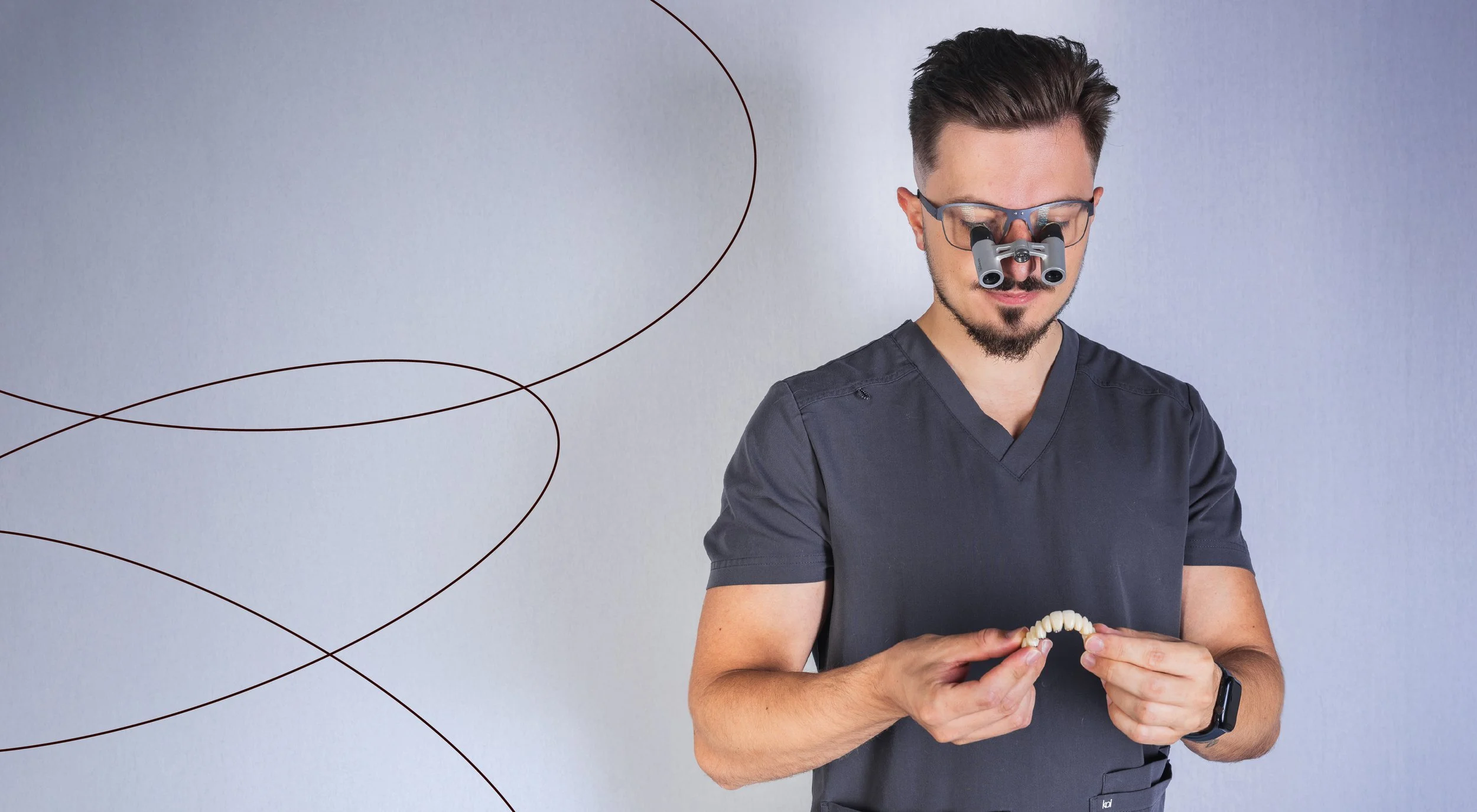 A man wearing glasses and a navy blue medical uniform uses binocular loupes to examine a dental model.