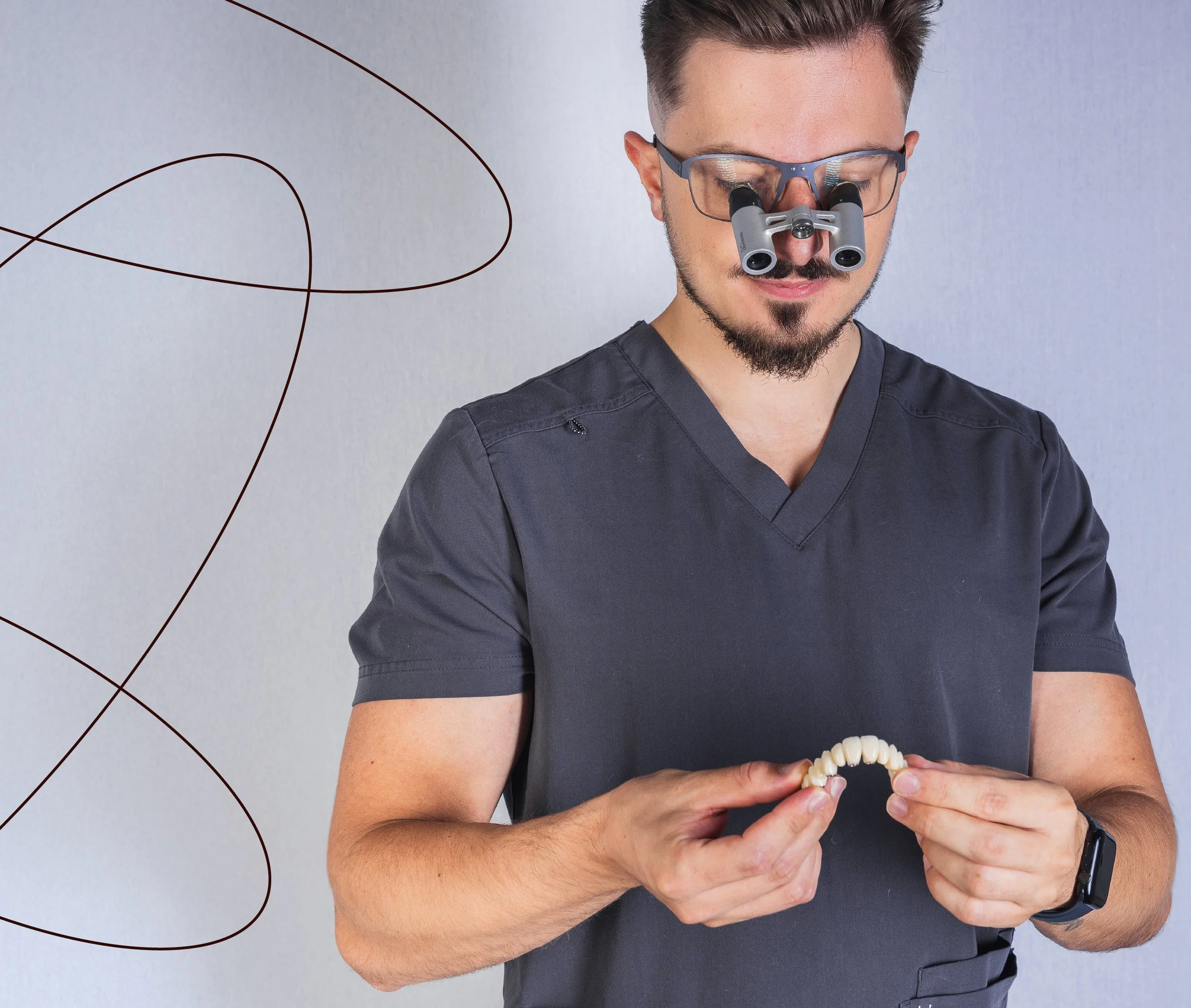 A male dentist wearing magnifying glasses with a dental loupe, holding a set of dental prosthetic teeth, standing against a plain background.
