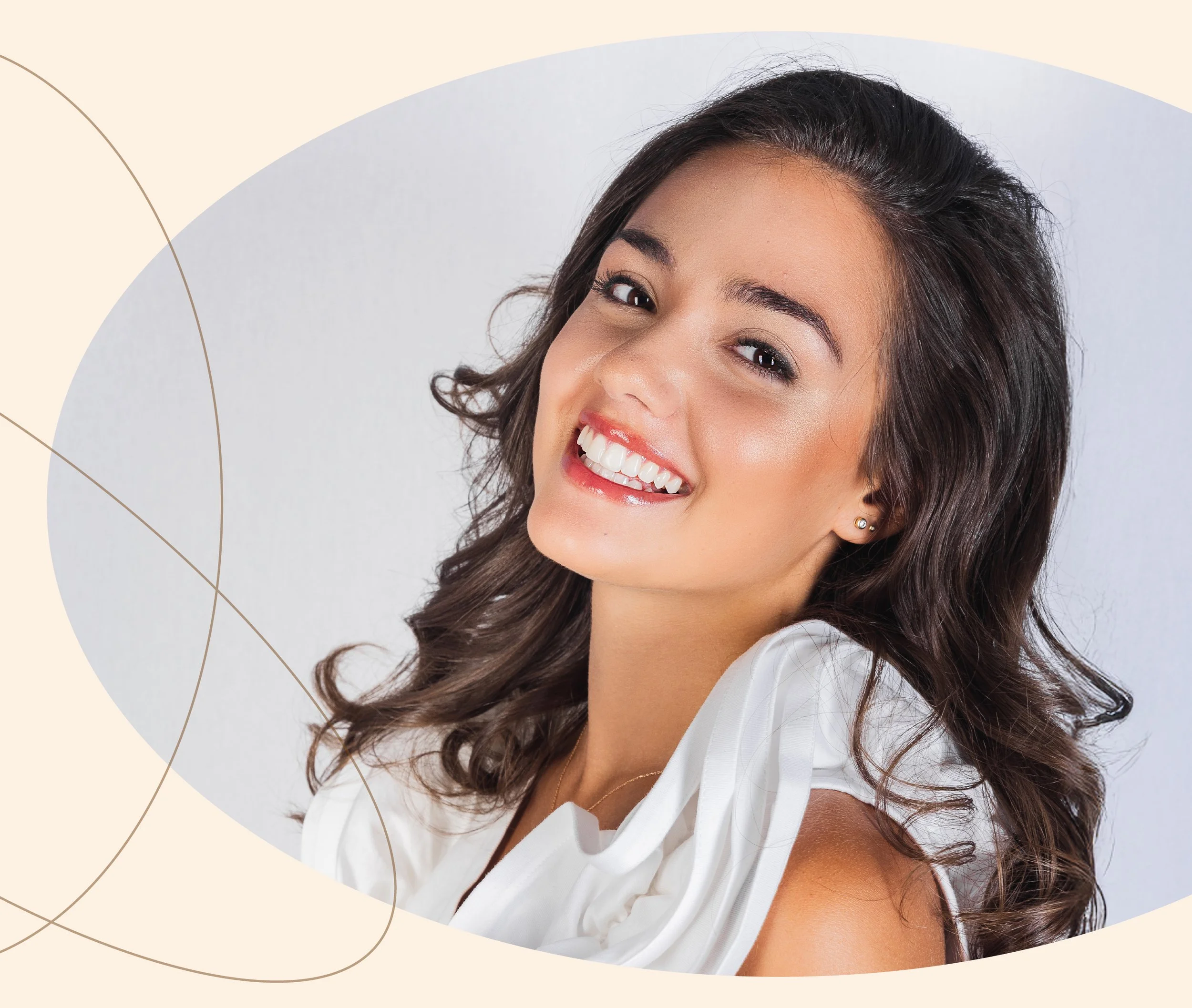 Close-up portrait of a smiling woman with dark wavy hair and earrings, wearing a white top against a light gray background.