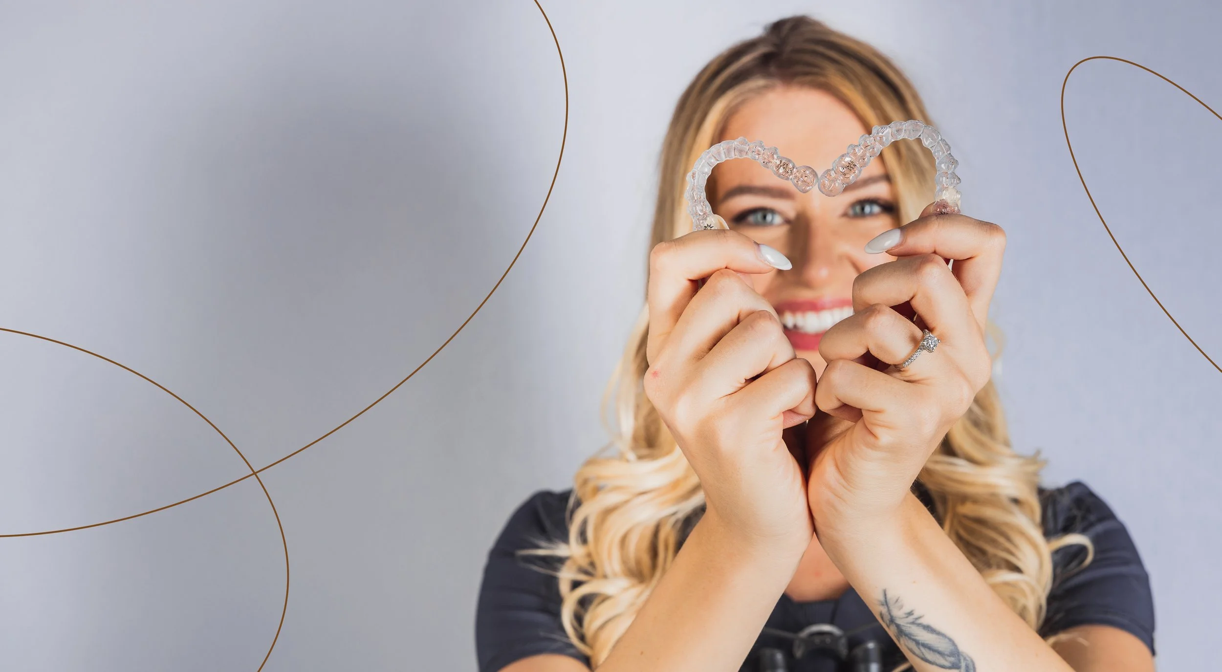 A smiling woman holding a clear, heart-shaped dental tray up to her face, with a light gray background and gold curved line decorations.