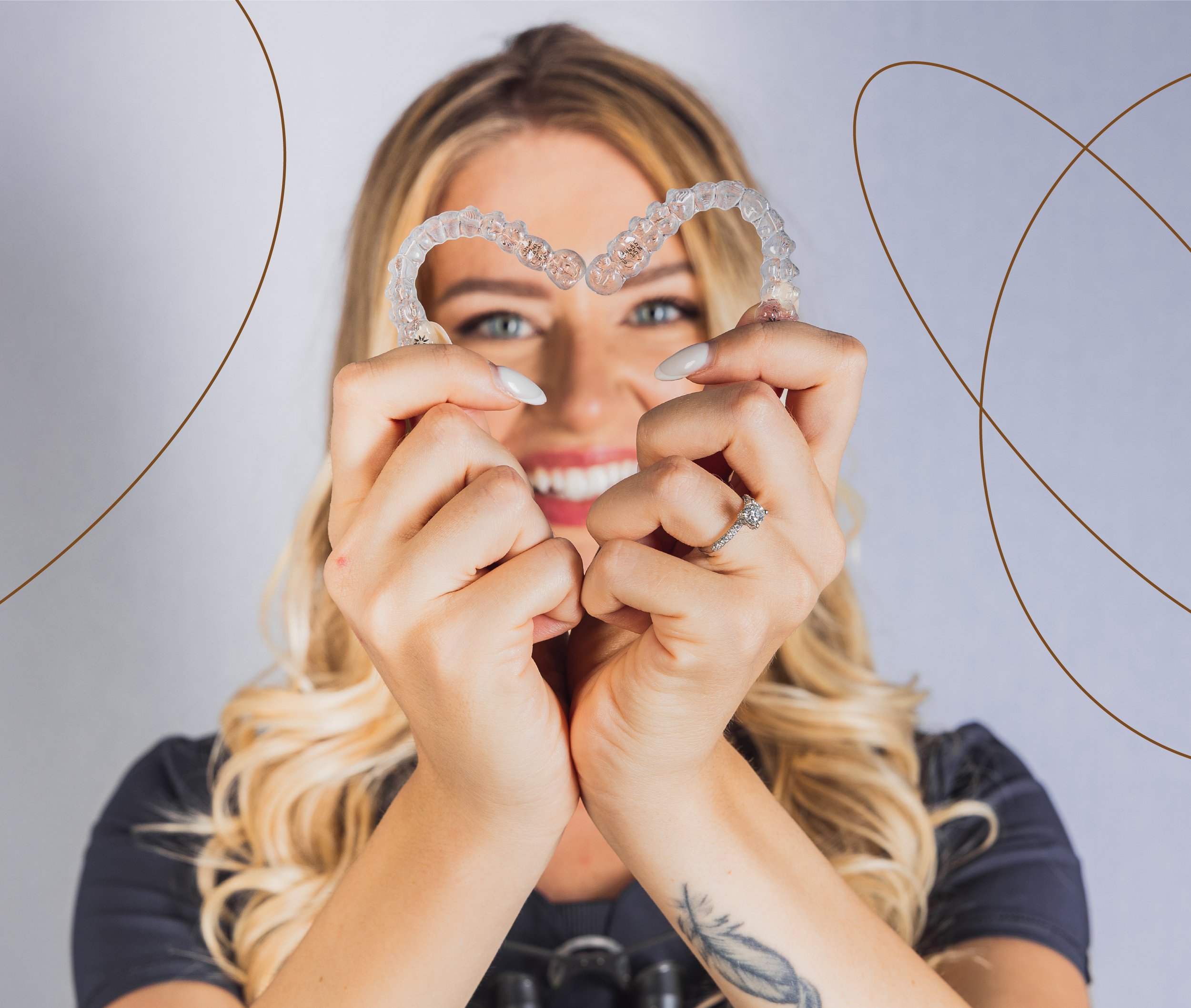 A woman with blonde hair holding a clear dental aligner shaped like a heart in front of her face, smiling.