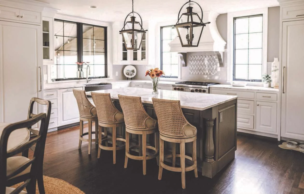 A bright kitchen with white cabinets, a large marble island, and five beige wicker barstools. There are two black-framed windows, a stove with a decorative white range hood, and a vase of orange flowers on the island. Two pendant lights hang above the island.