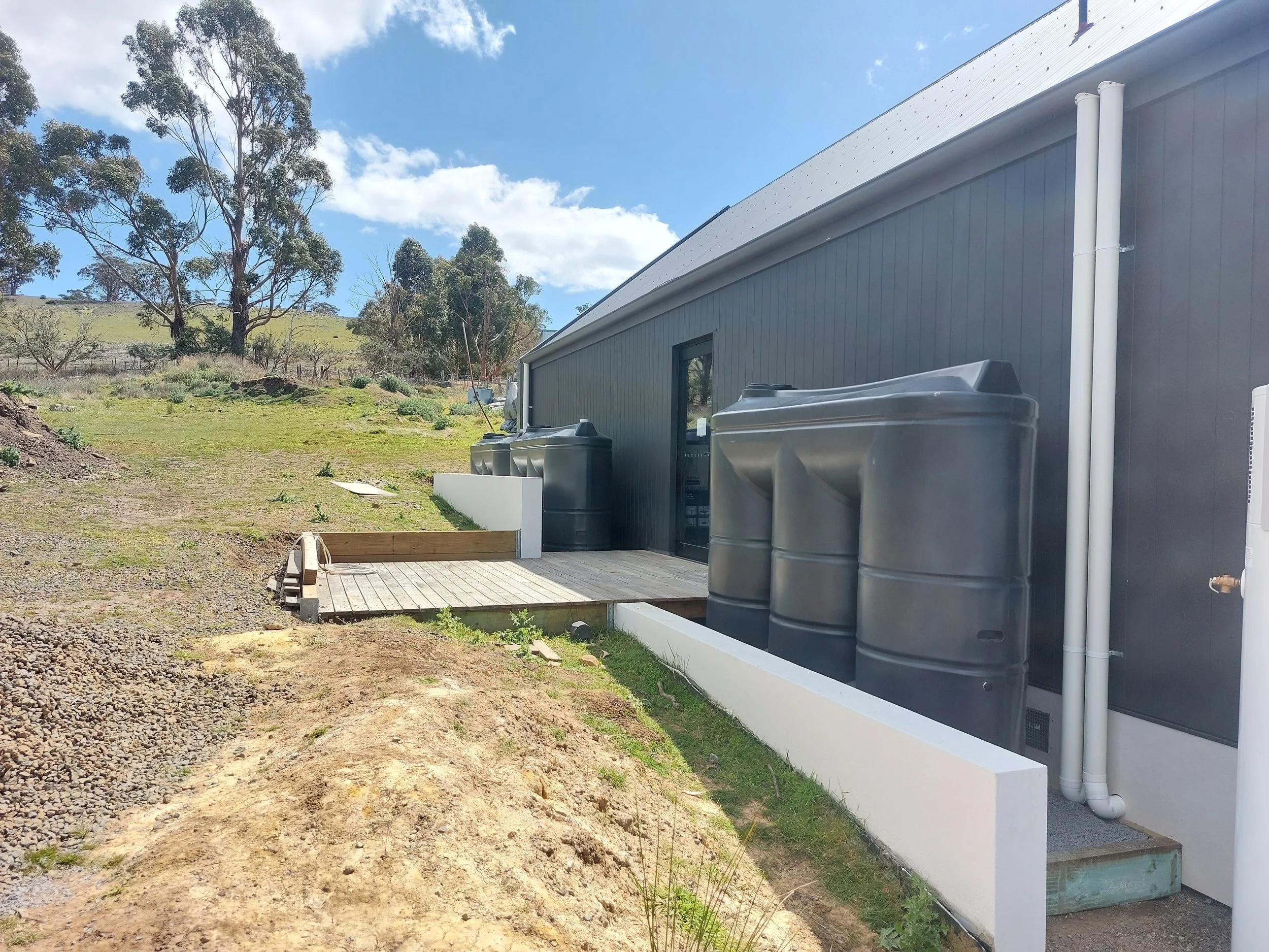 Side view of a modern gray building with large black water tanks, white piping, and a small wooden deck on a grassy hillside under a partly cloudy sky.