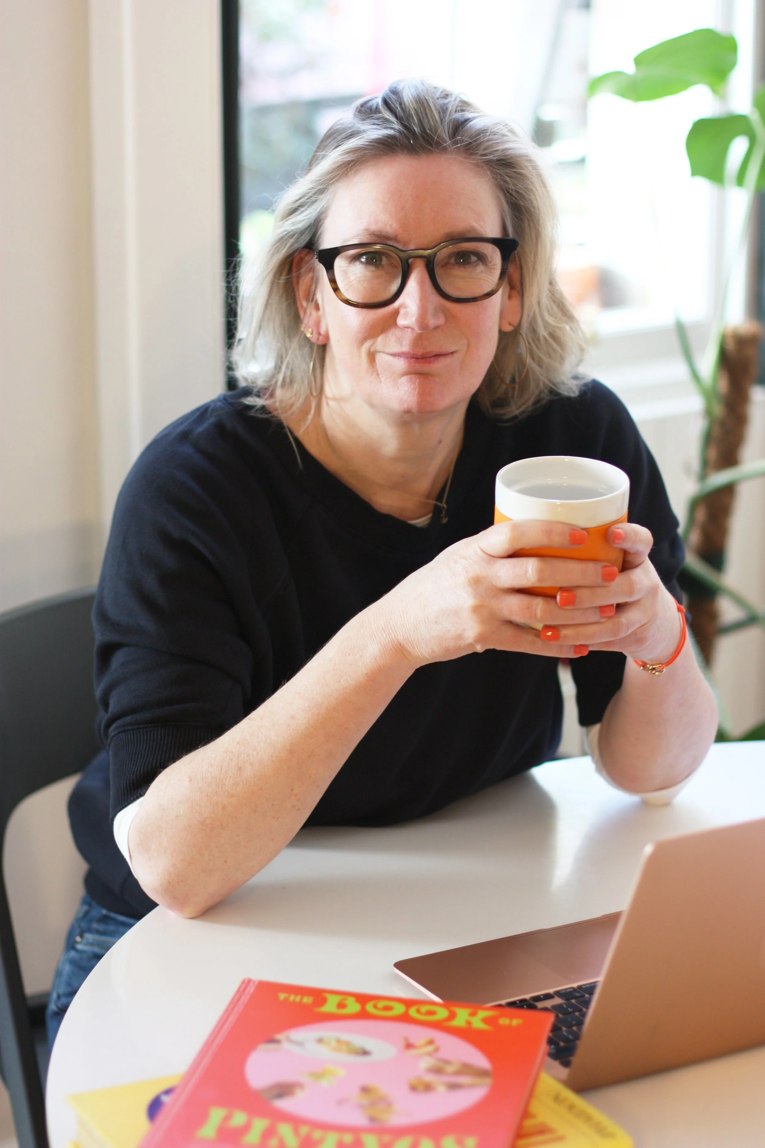 Photo of Rachel Athey, founder, with glasses and shoulder-length hair sitting at a table, holding a cup with both hands, smiling slightly, with a laptop and a red book of pintxo recipes nearby.