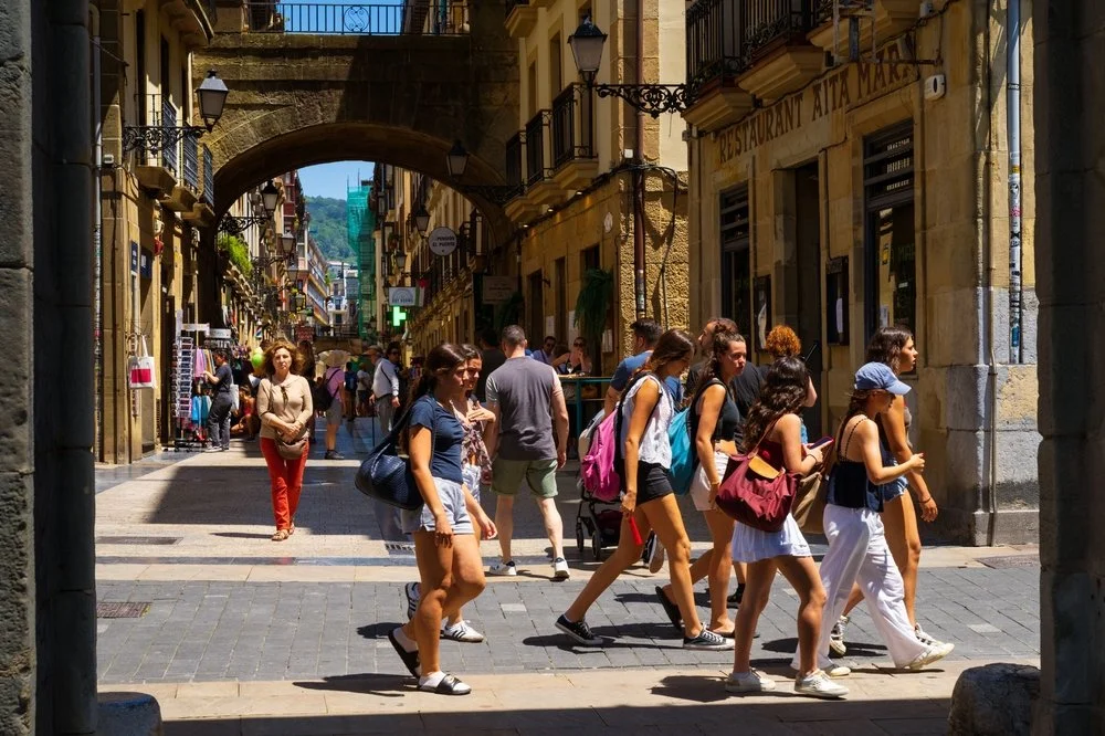 Teenagers walking on a cobblestone street in a European city with colorful buildings and shop signs, under a stone archway with hillside in the background.