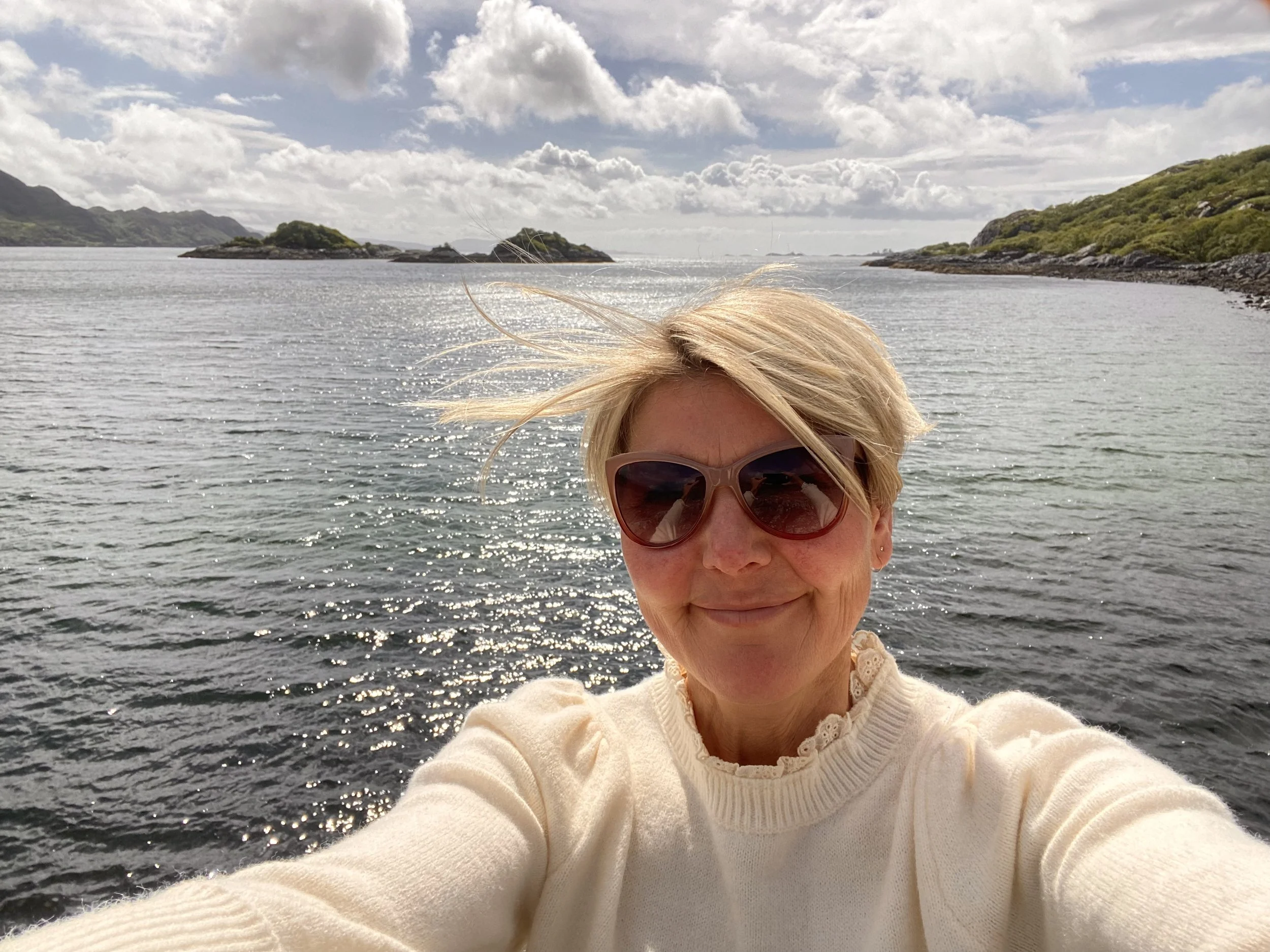 A woman with blonde hair wearing sunglasses and a cream-colored sweater taking a selfie by a body of water with islands and hills in the background.